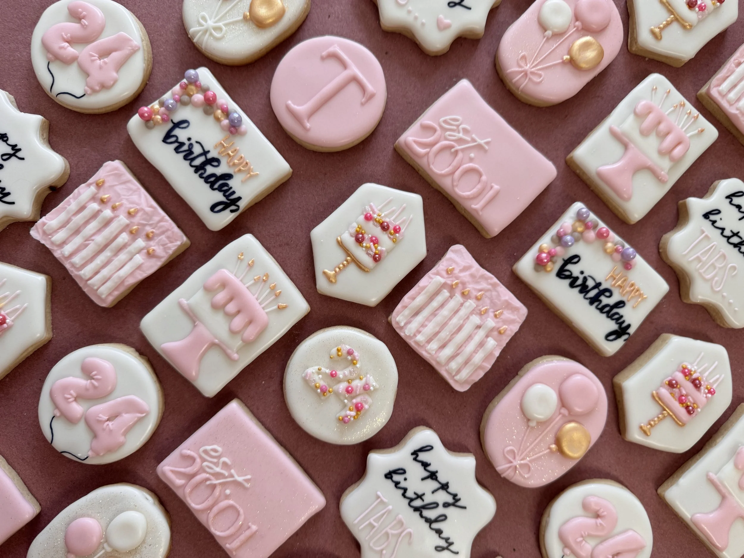 Decorated cookies with pink and white icing, some with birthday cake designs, balloons, and the words "Happy Birthday" and "24th" written on them, arranged on a pink surface.
