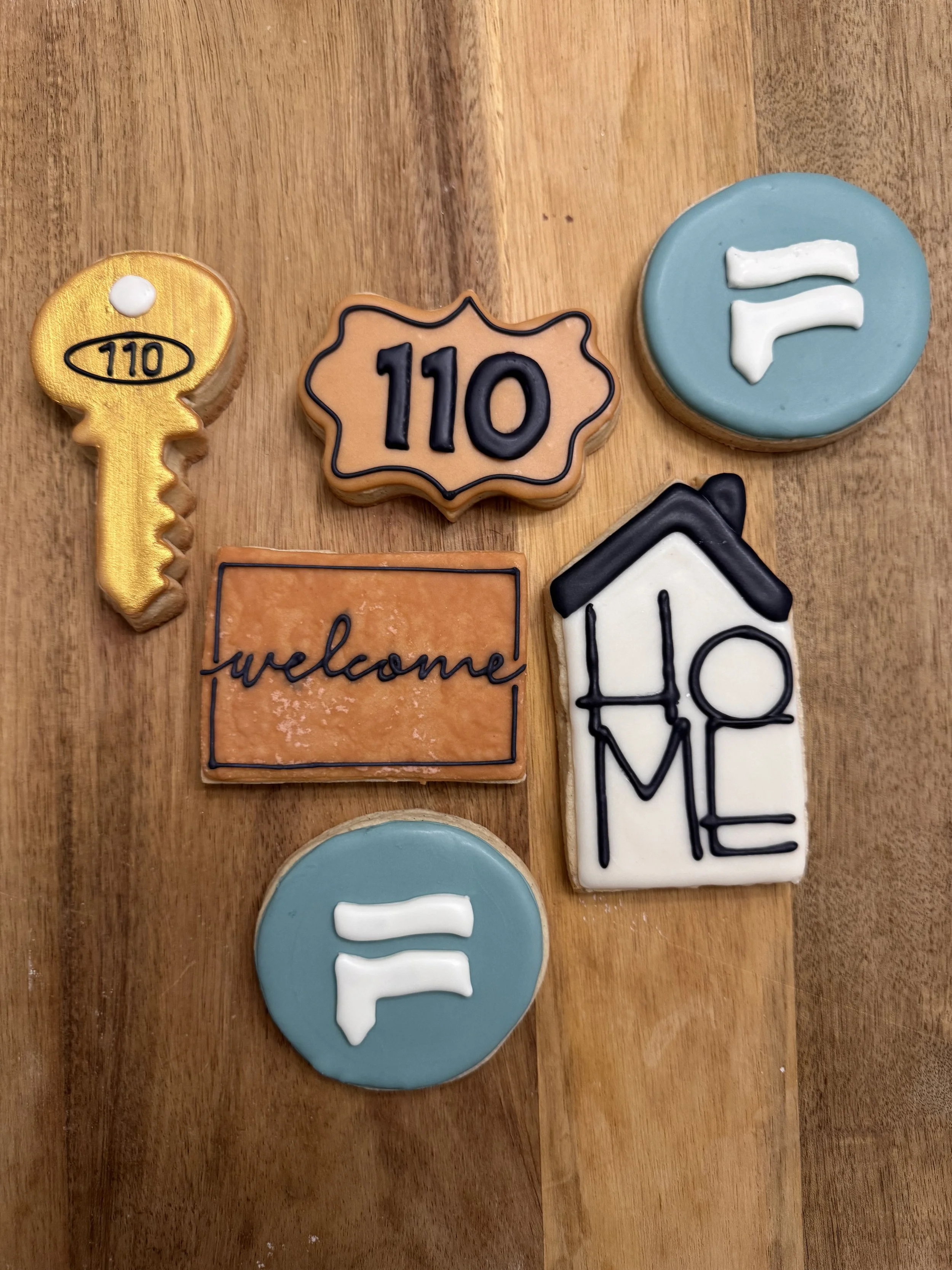 Decorative cookies on a wooden surface, including a key, a HOUSE sign with '110', a blue circle with 'F', a rectangular cookie with 'welcome' written, and a house-shaped cookie with 'HOME' written.