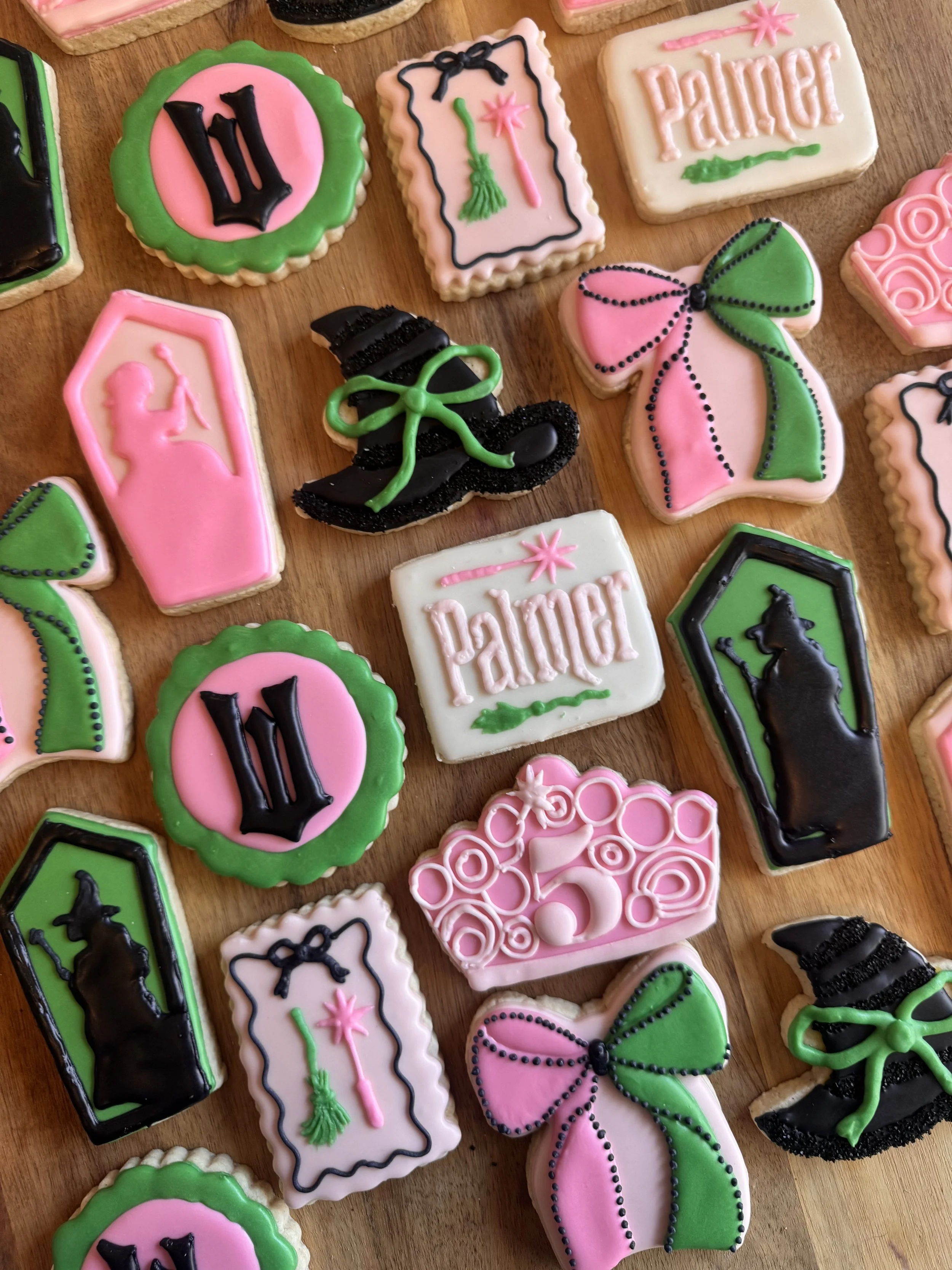 Decorated sugar cookies with pink, green, black, and white icing, featuring decorative designs such as bows, witch hats, the word 'Palmer', and silhouettes of cats, all arranged on a wooden surface.