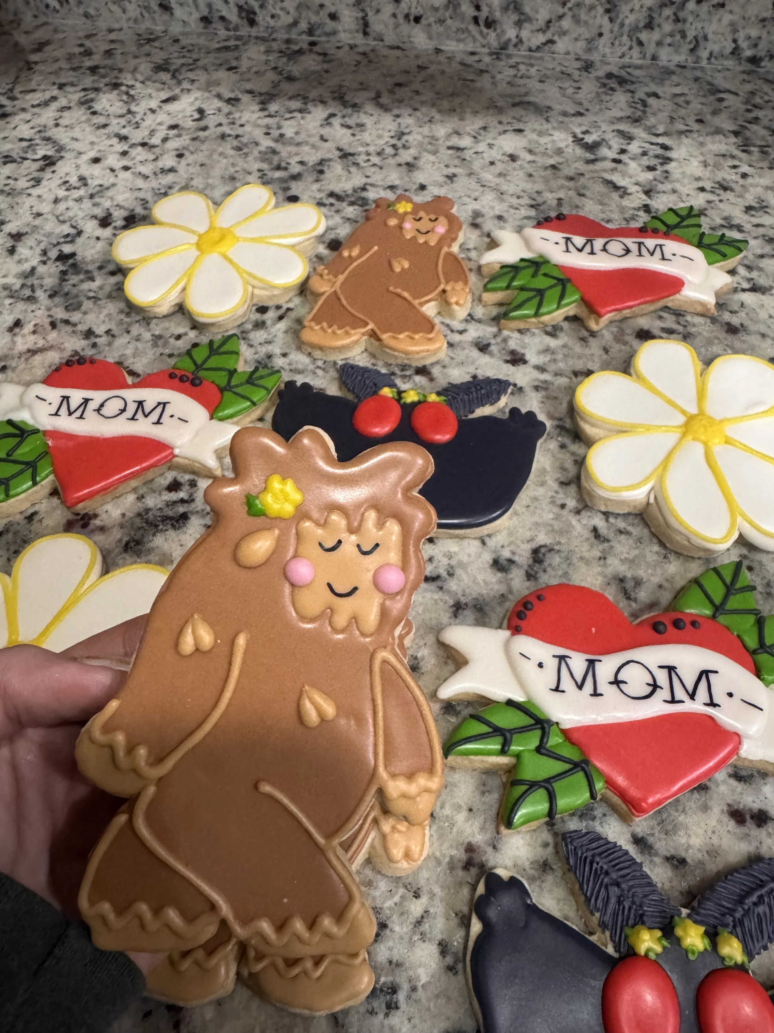 Decorative cookies placed on a granite countertop, including a Gnome, flowers, hearts with 'MOM' written, and holly leaves with berries.