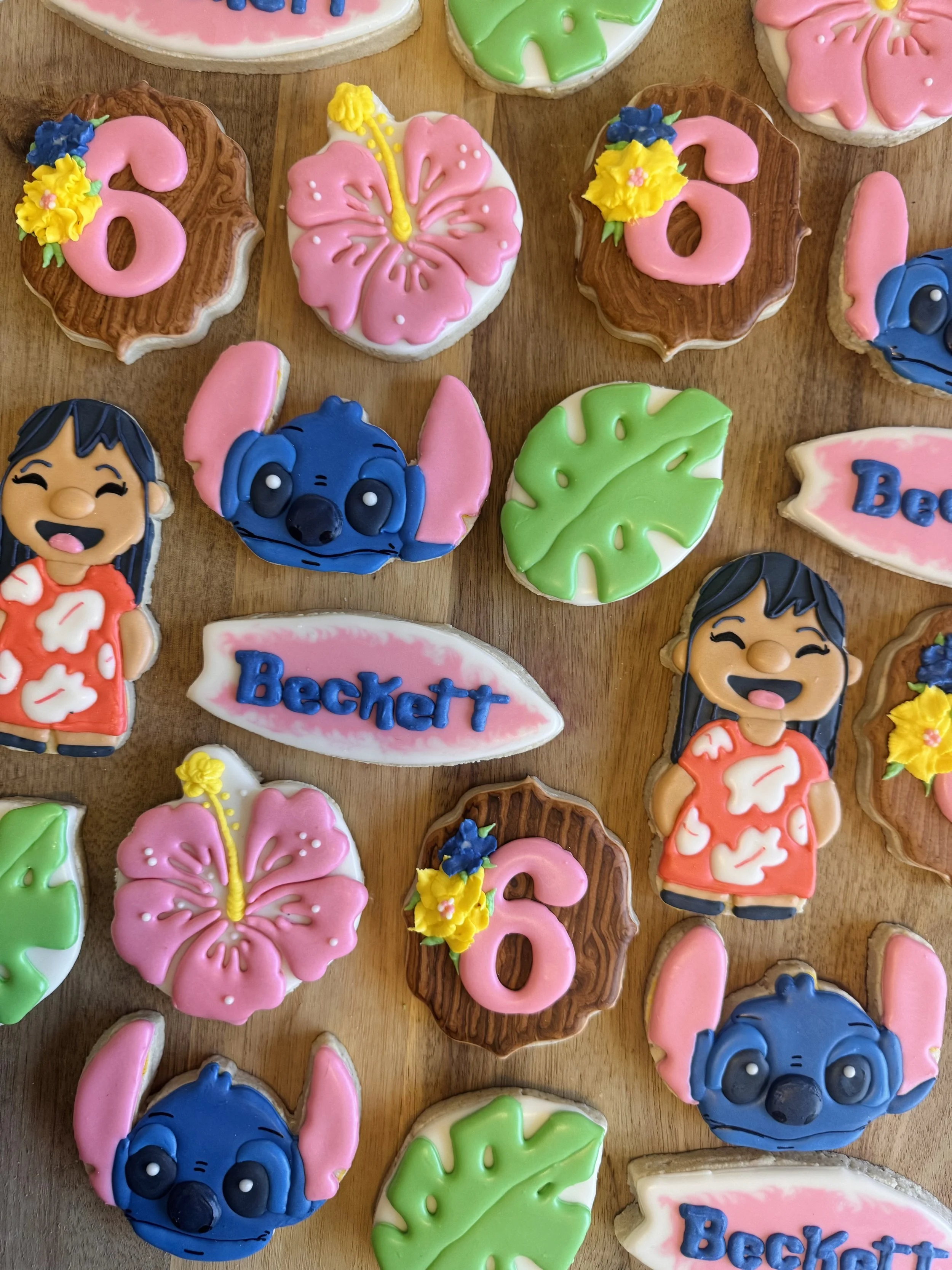 Decorative cookies with tropical theme, including pink hibiscus flowers, green monstera leaves, blue Stitch character, a girl in a red floral dress, and signs with the name "Beckett".