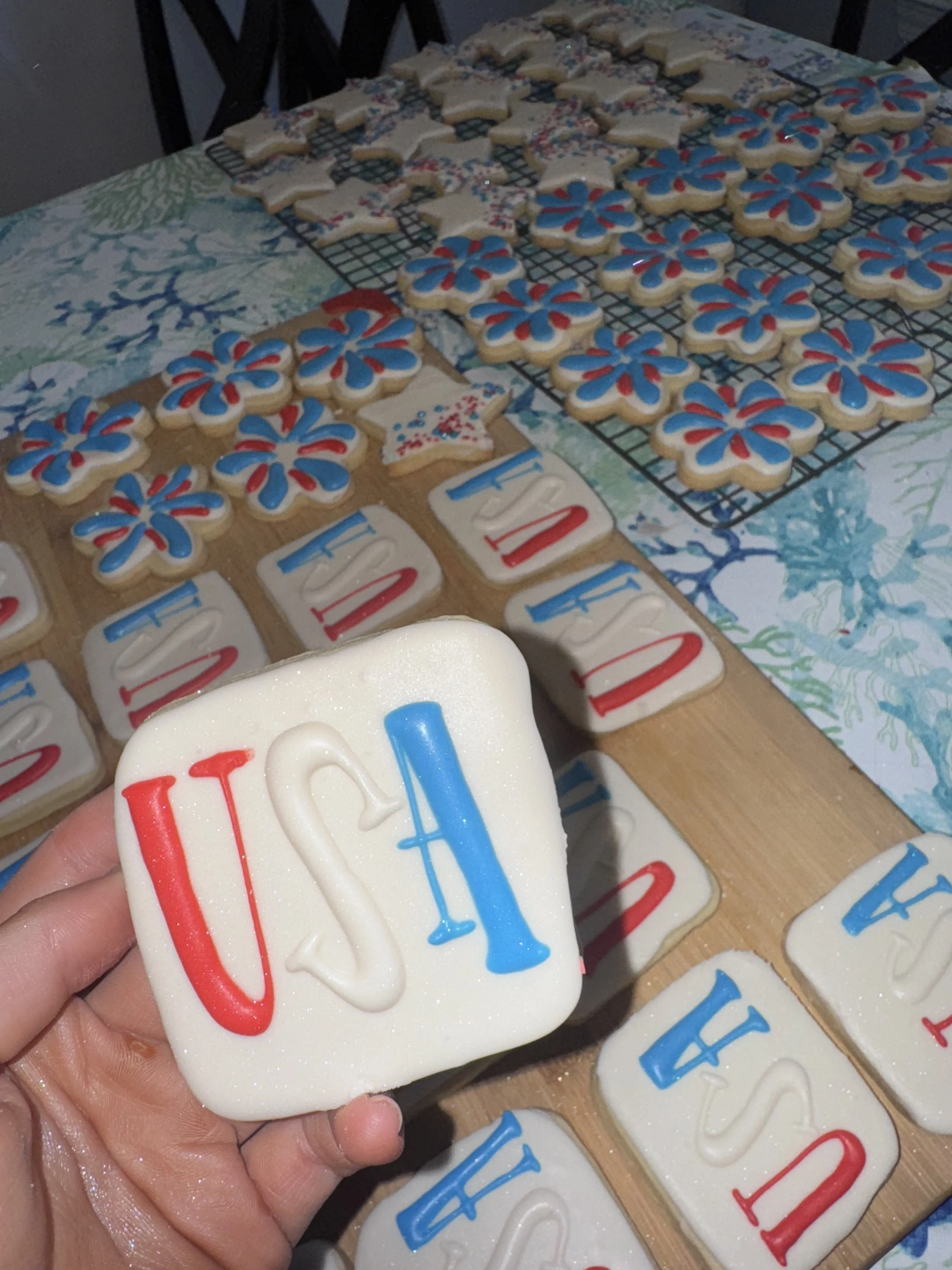 Decorated sugar cookies with patriotic red, white, and blue icing, spelling out 'USA' and featuring fireworks and stars, laid out on a table and cooling rack, likely for a Fourth of July celebration.