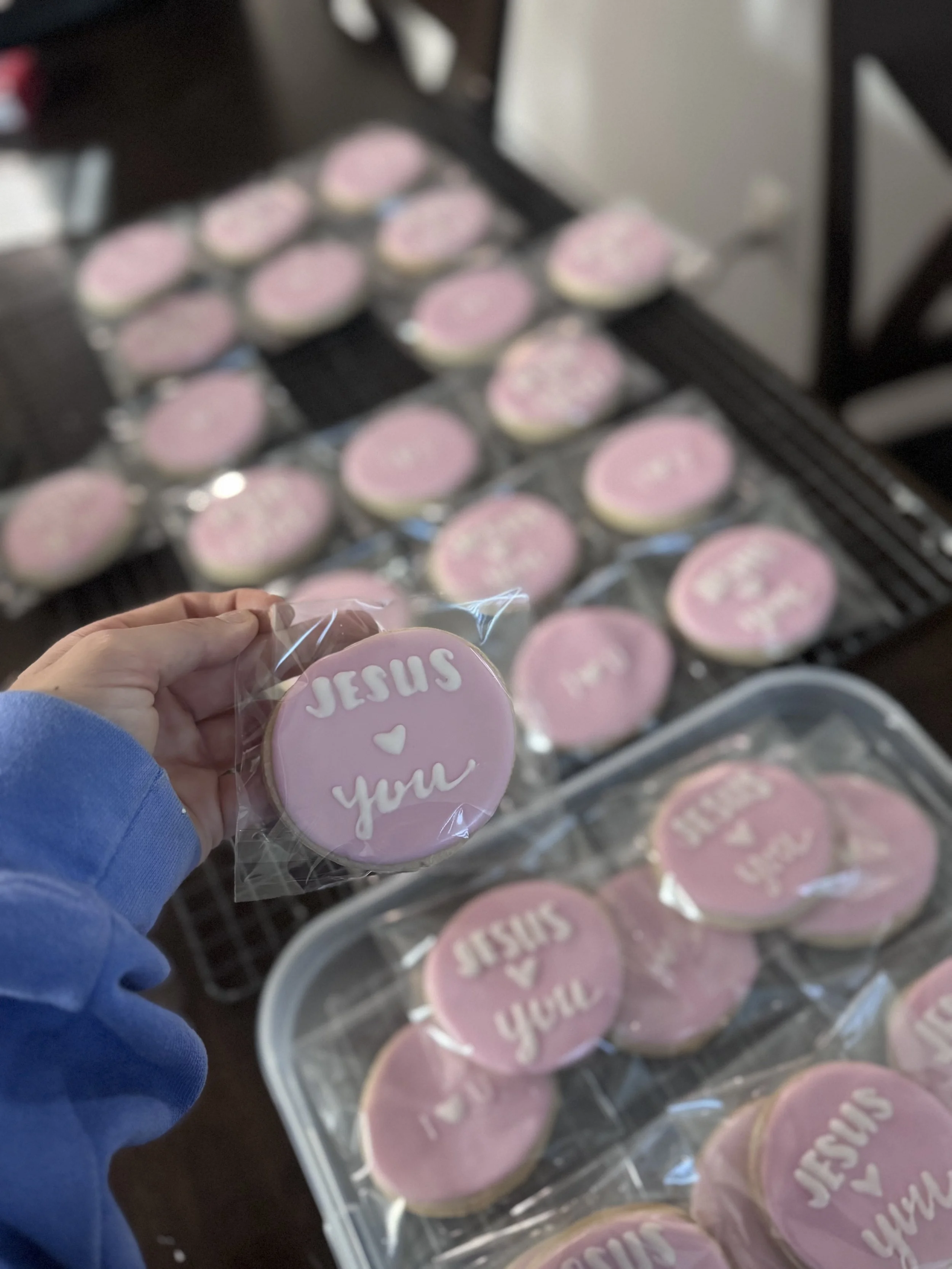 Pink decorated cookies with the words 'Jesus ❤️ you' written in white icing, some of which are individually wrapped.