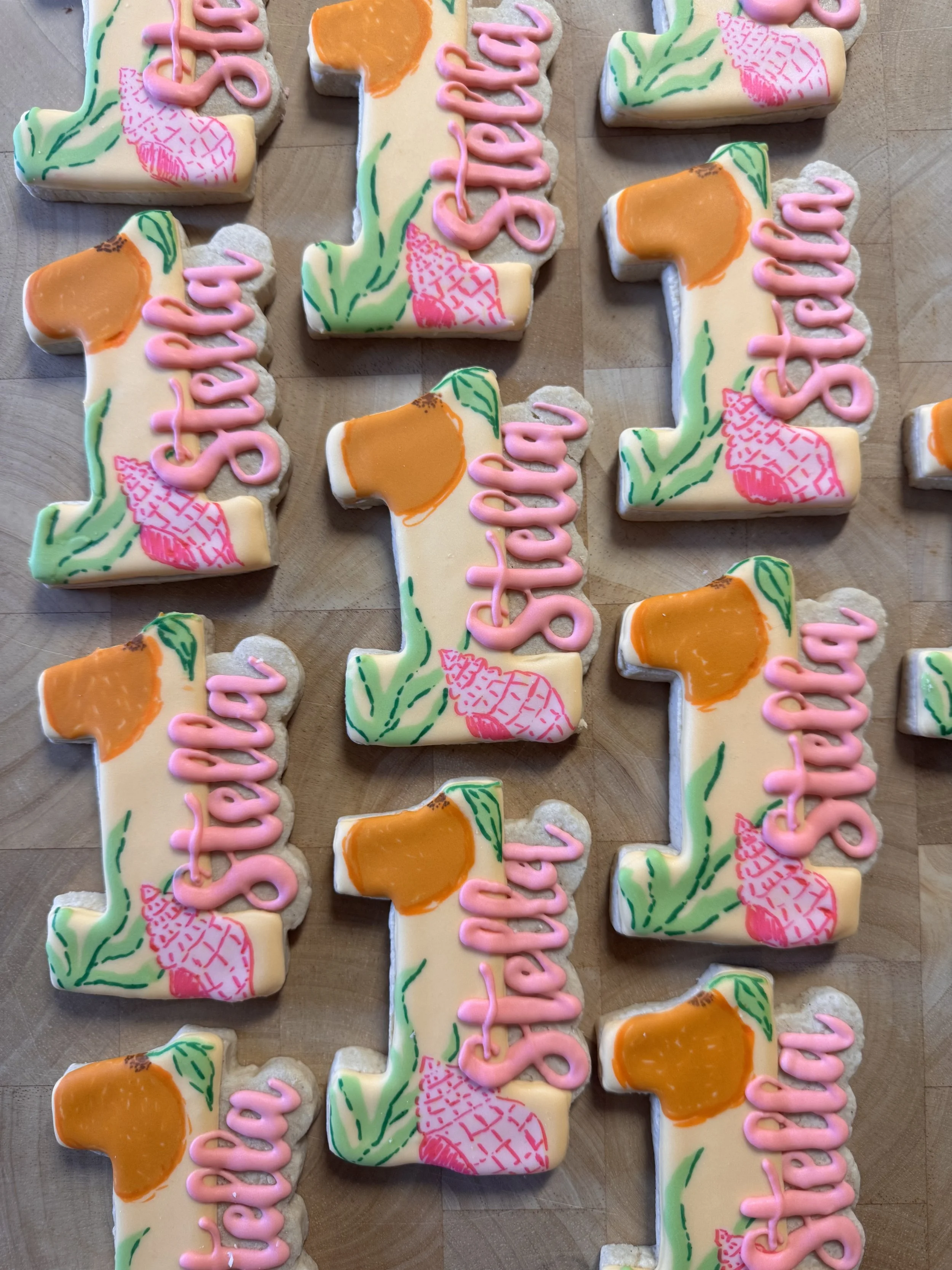Cookies decorated with the phrase  'Stella," a seashell, some seaweed, and a large orange slice, featuring colorful icing and pink script.
