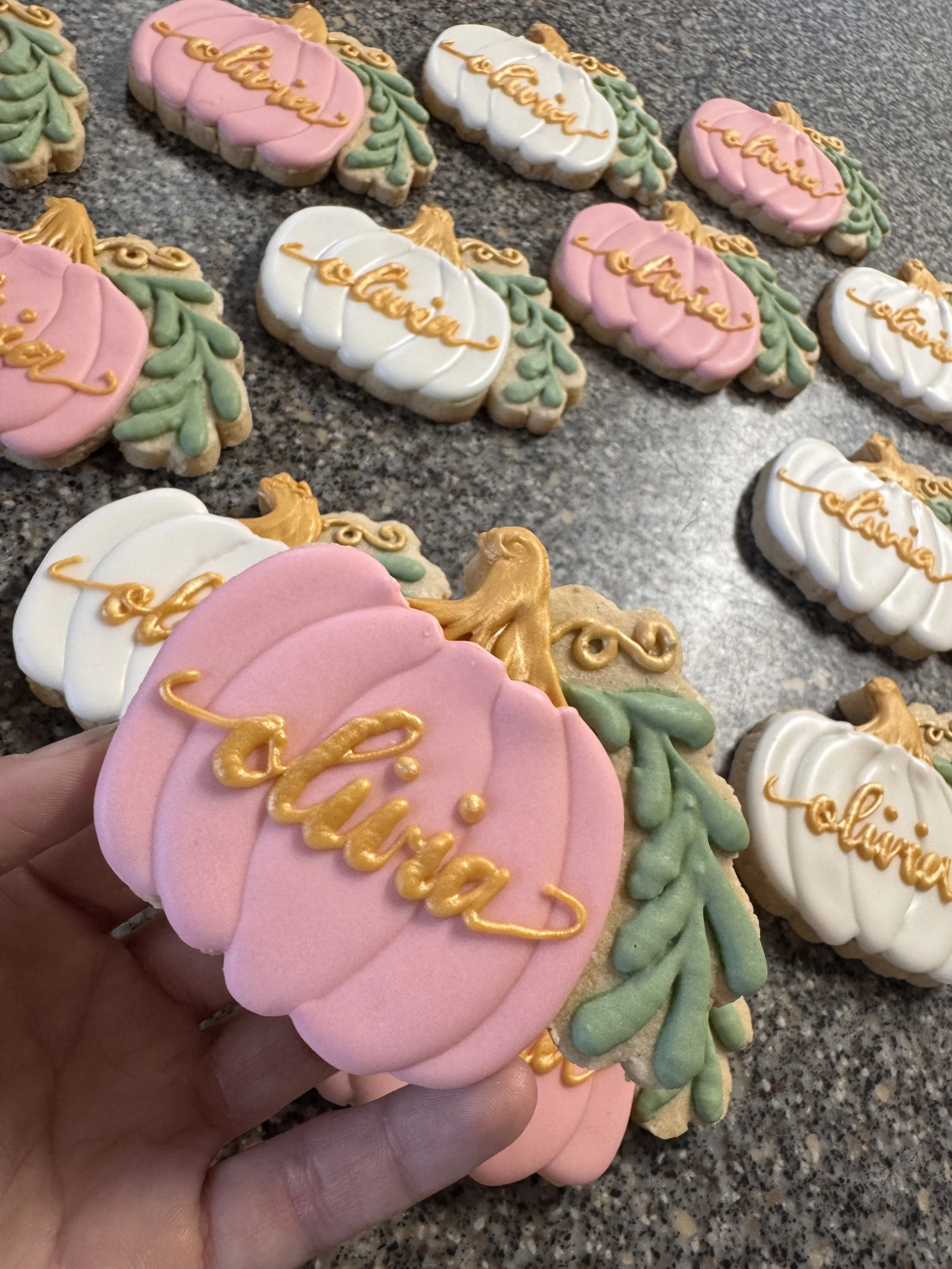 Assorted decorated sugar cookies shaped like pumpkins with pink and white icing, green leaves, and the word 'olivia' written in gold icing.