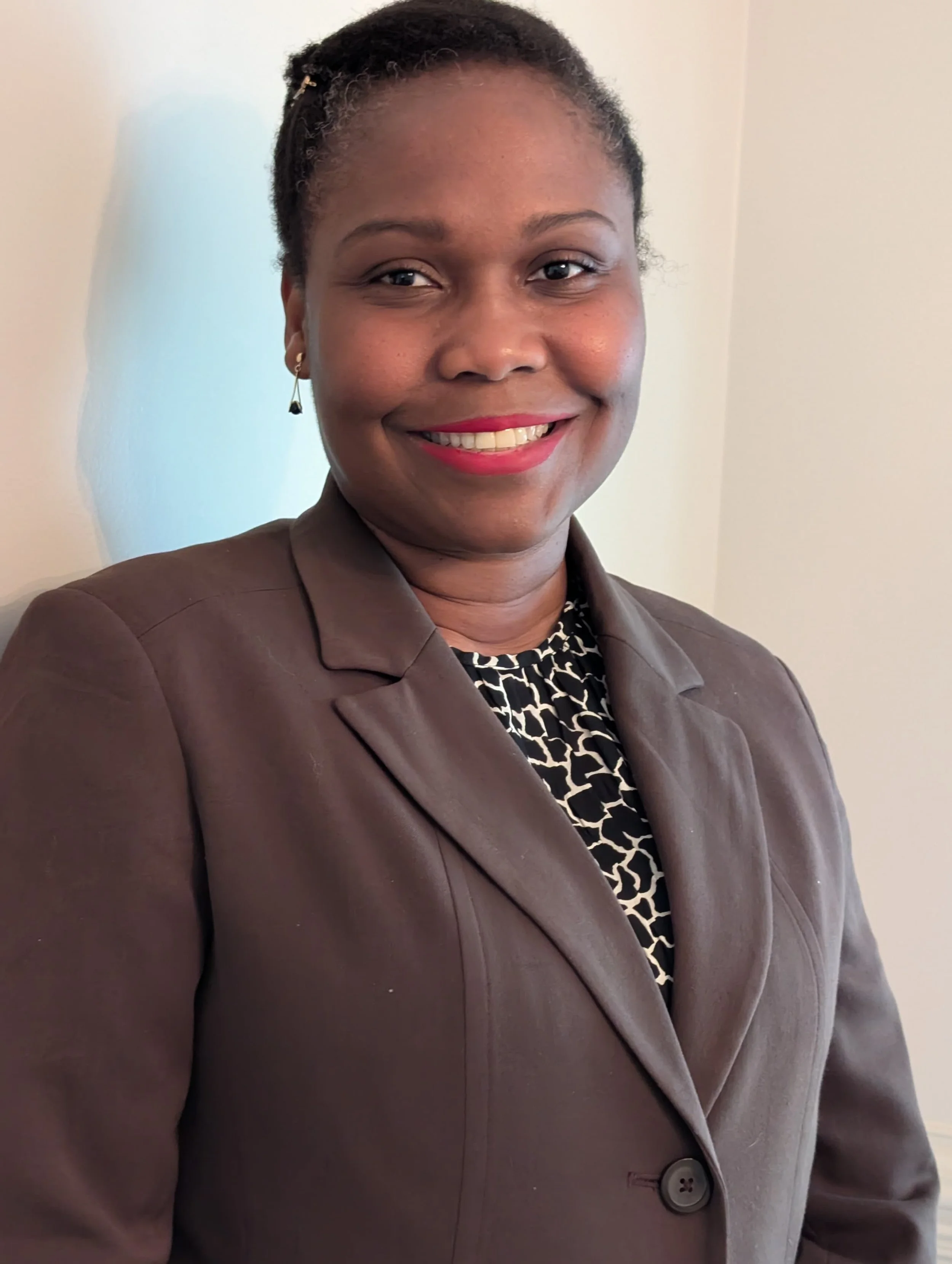 A smiling woman dressed in a brown blazer and a black and white patterned top, standing against a plain wall.