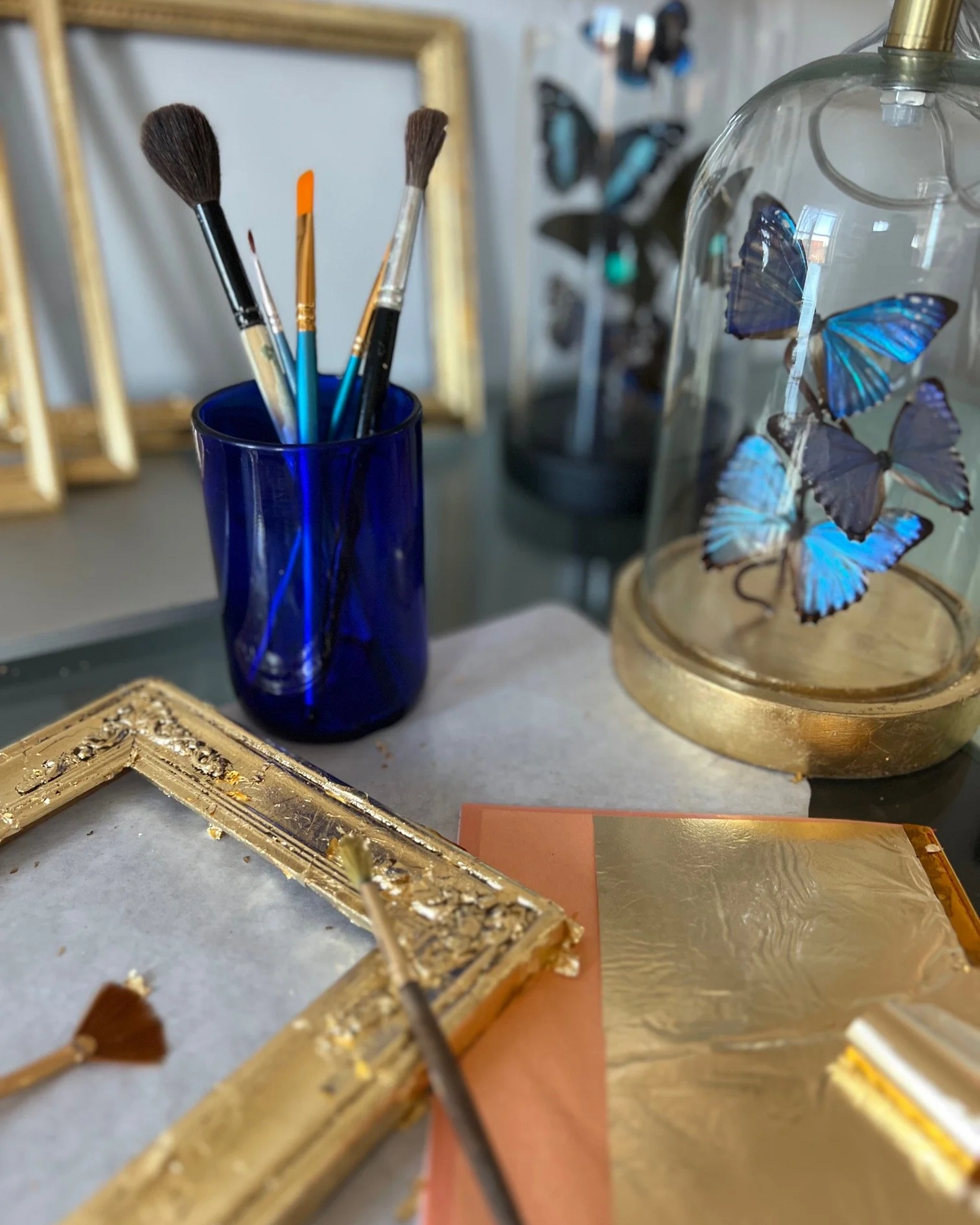 A desk with a blue glass container holding paintbrushes, a decorative glass dome with blue butterflies, a gold picture frame, and gold spray paint on a sheet of paper.