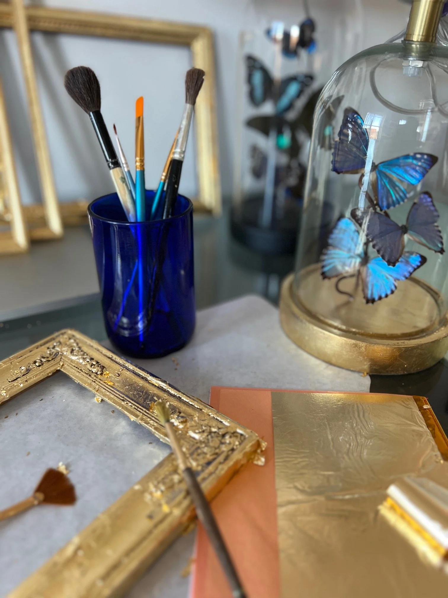 Art and craft supplies including paintbrushes in a blue glass container, a decorated glass dome with blue butterfly decorations, a gold ornate picture frame, a small palette knife, and sheets of gold foil on a table.