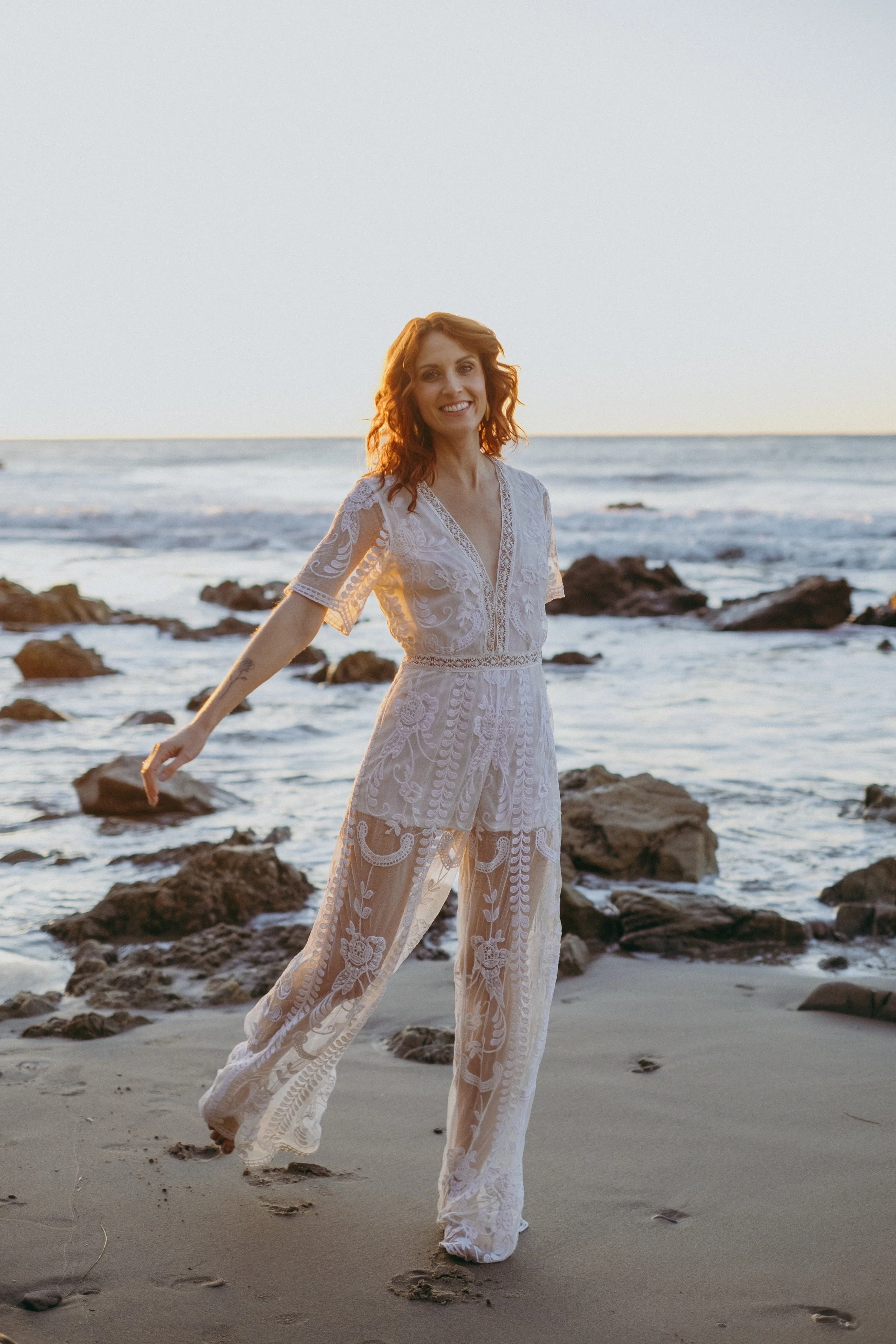 A woman in a white lace jumpsuit stands on a beach with rocks, smiling at sunset with ocean in the background.