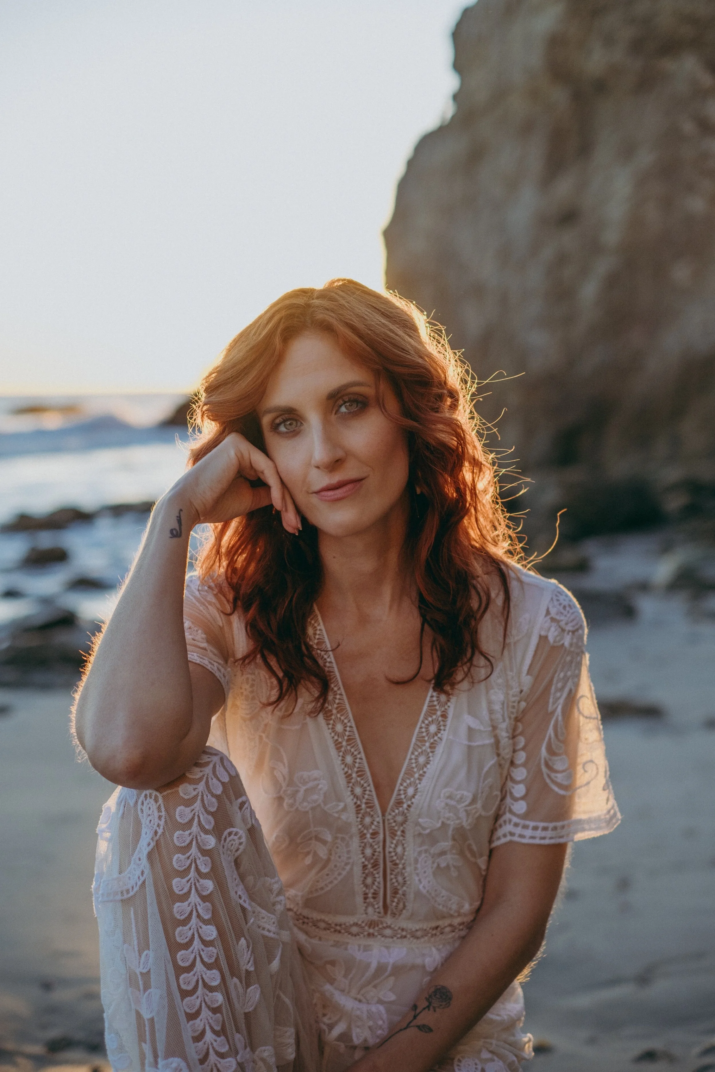 A woman with wavy red hair and tattoos is sitting on a beach during sunset, wearing a lace dress, with a rocky cliff in the background.