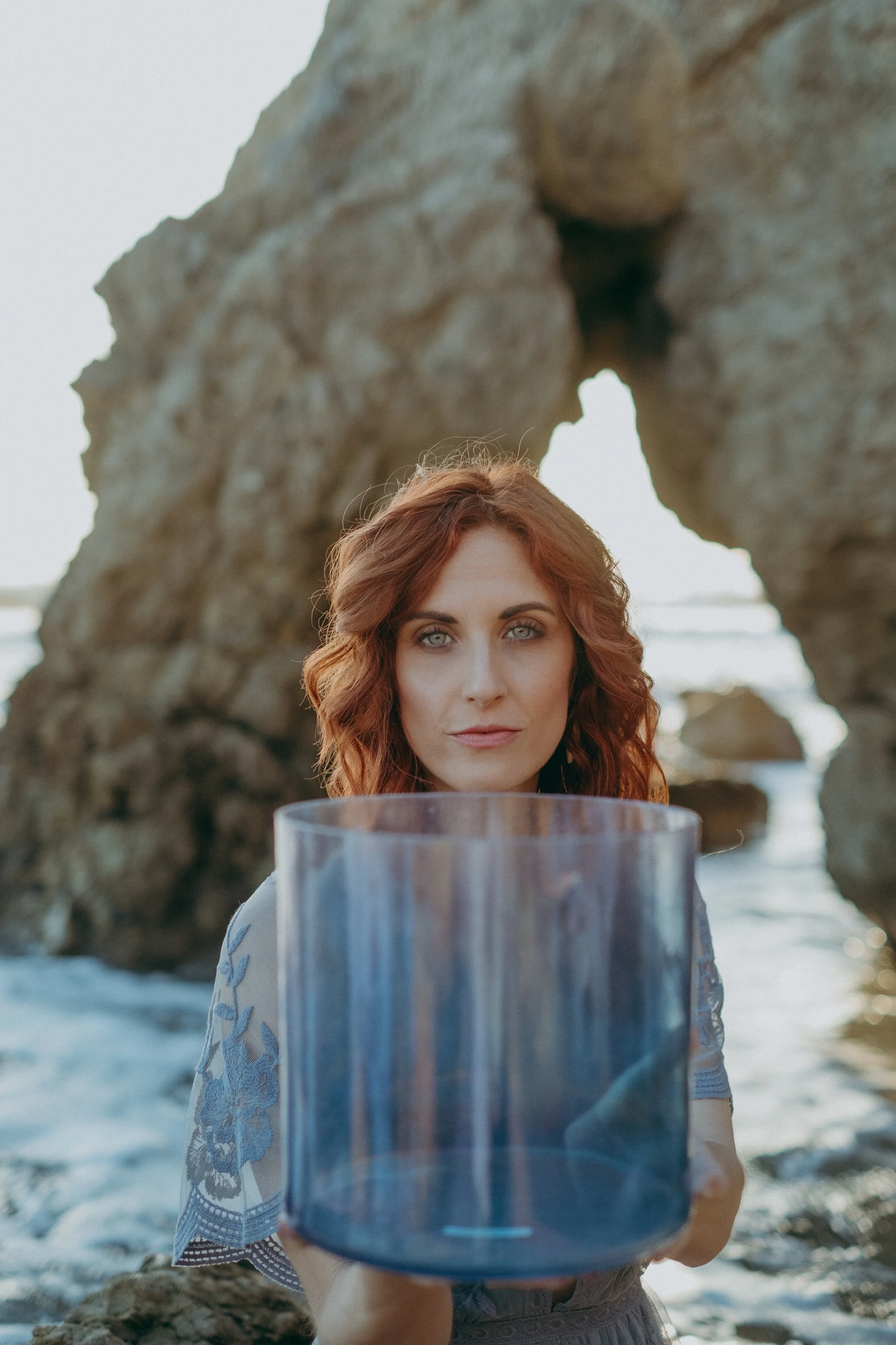 A woman with red hair and blue eyes holding a transparent blue bucket at the beach with large rocks and ocean in the background.