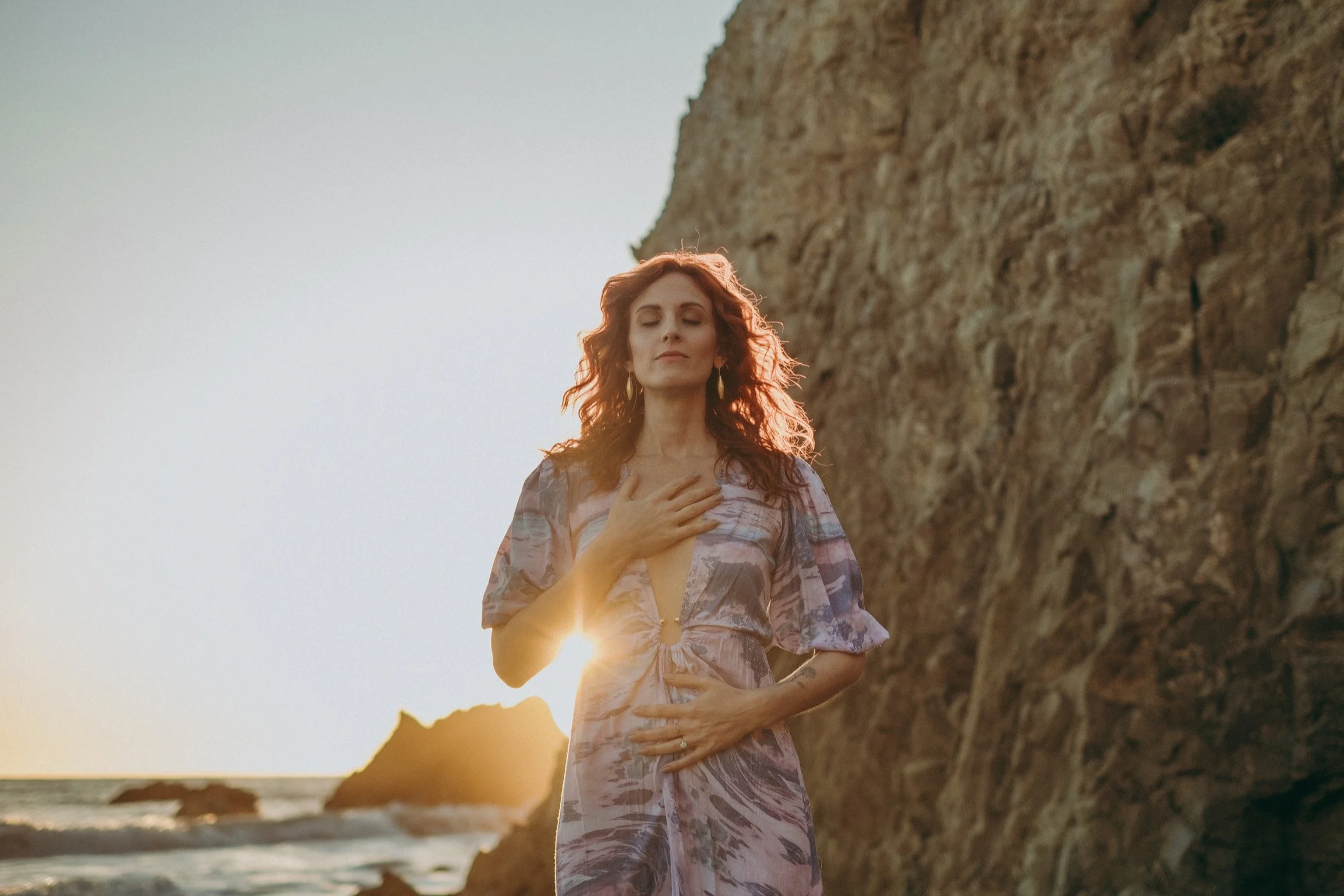 A woman standing on a beach at sunset with her eyes closed, wearing a floral dress, with one hand on her chest and the other on her stomach, near a large rocky cliff.