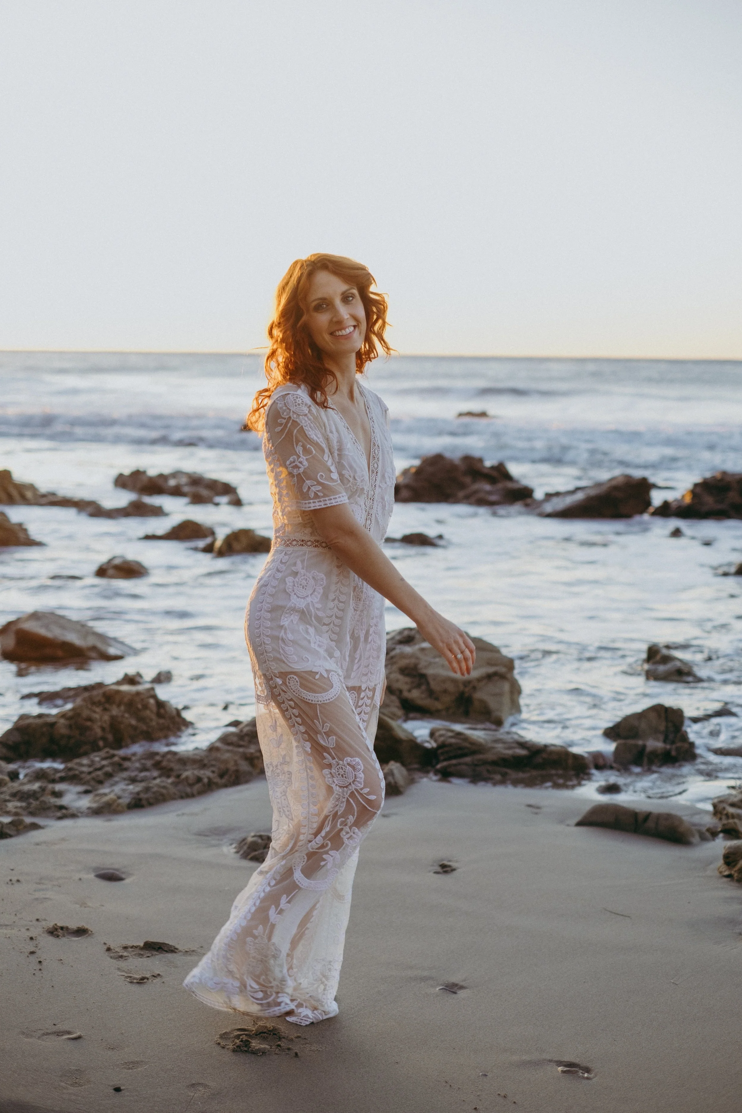 A woman in a white lace dress walking on the beach with rocks and the ocean in the background at sunset.