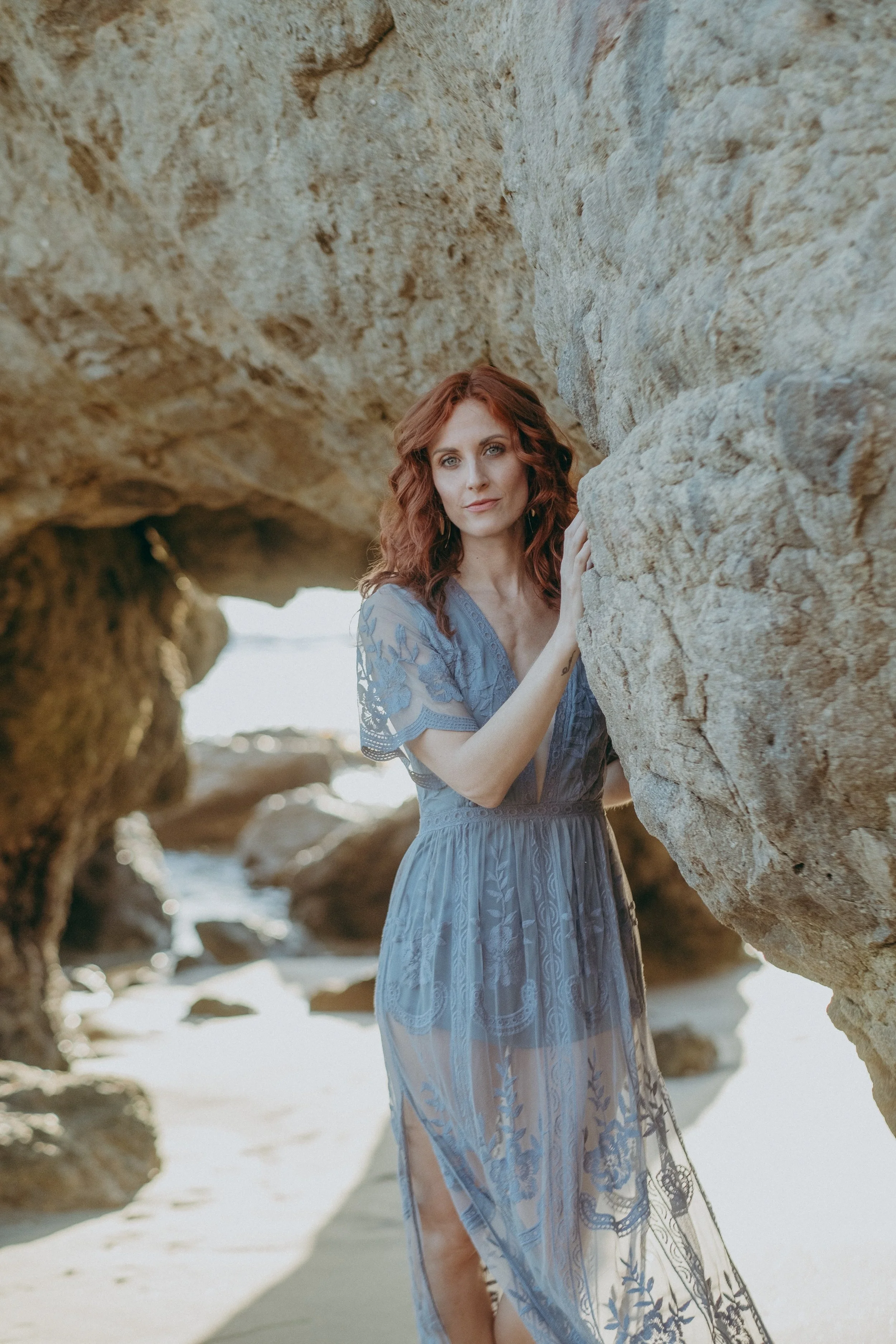Woman with red hair standing on a beach, peeking through large rocks in a sheer blue lace dress.
