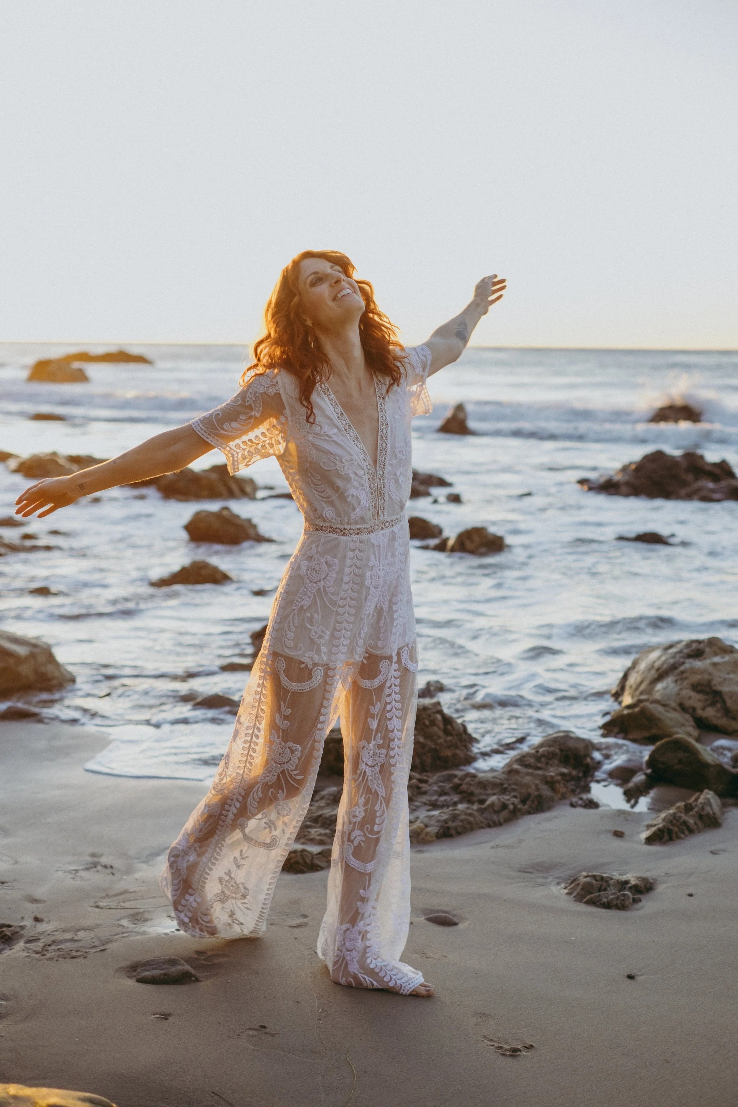 A woman with red hair in a white lace dress standing on a rocky beach with arms outstretched and facing the sunset.