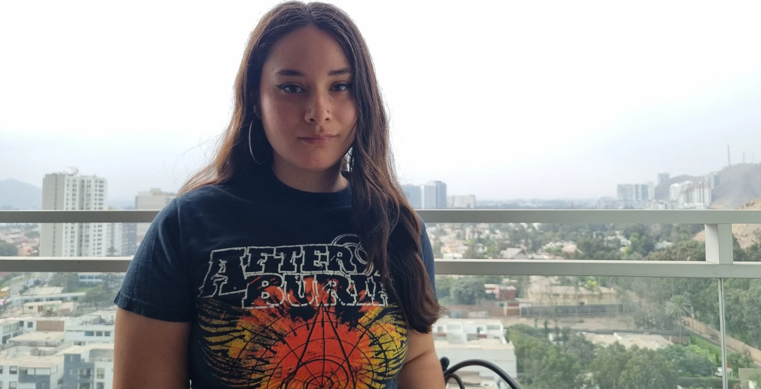 A young woman with long brown hair stands on a balcony with city buildings and hills in the background, wearing a black graphic T-shirt from a death metal, deathcore, metalcore band After The Burial.