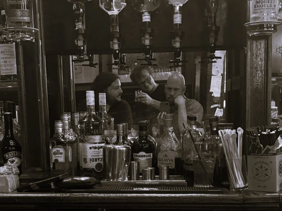 Three men behind a bar with various bottles of alcohol, mirrors, and bar equipment, taking a selfie.