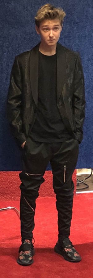 Young male with blonde hair wearing a black leather jacket, black t-shirt, black pants with zippers, and black sneakers with a floral pattern, standing against a dark blue wall on a red carpet.