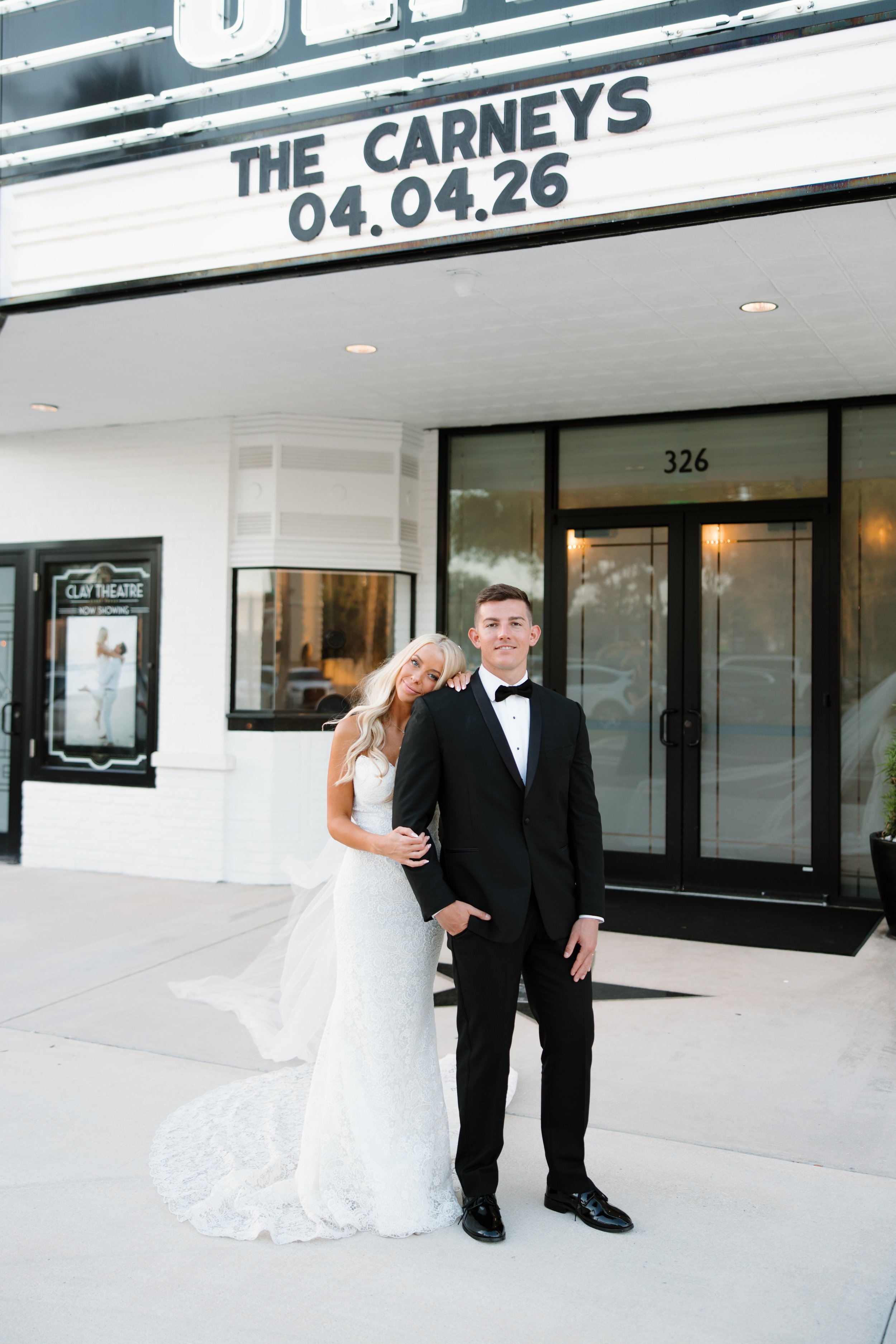 A couple dressed in wedding attire standing outside a theater building with a marquee sign that reads 'The Carneys 04.04.26'. The bride is in a white lace wedding gown with a long train, and the groom is in a black tuxedo with a bowtie. They are smil