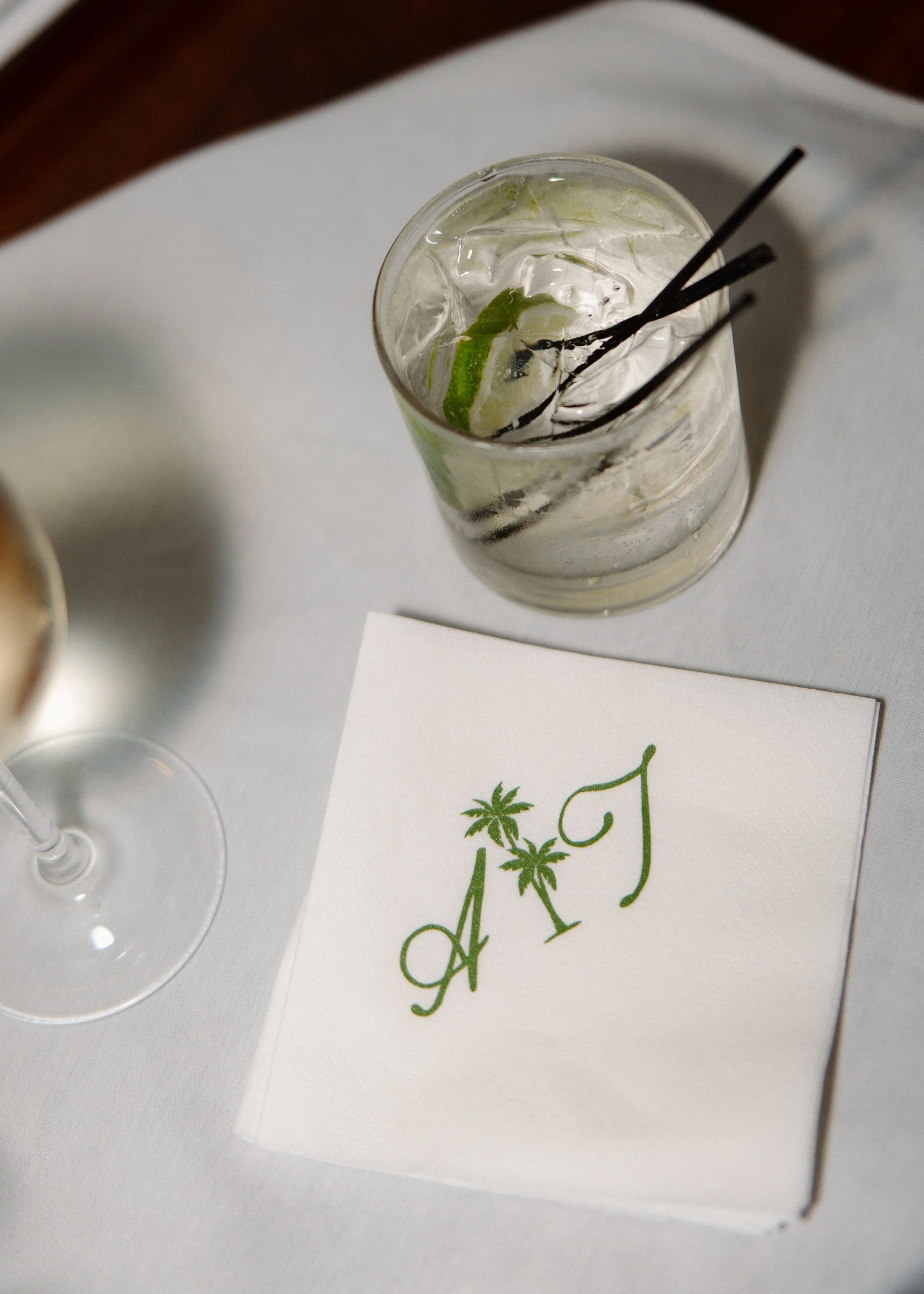A glass of clear beverage with lime wedge, ice, and black stir sticks on a white table. Part of a menu or card with green decorative writing and palm tree illustrations is visible.