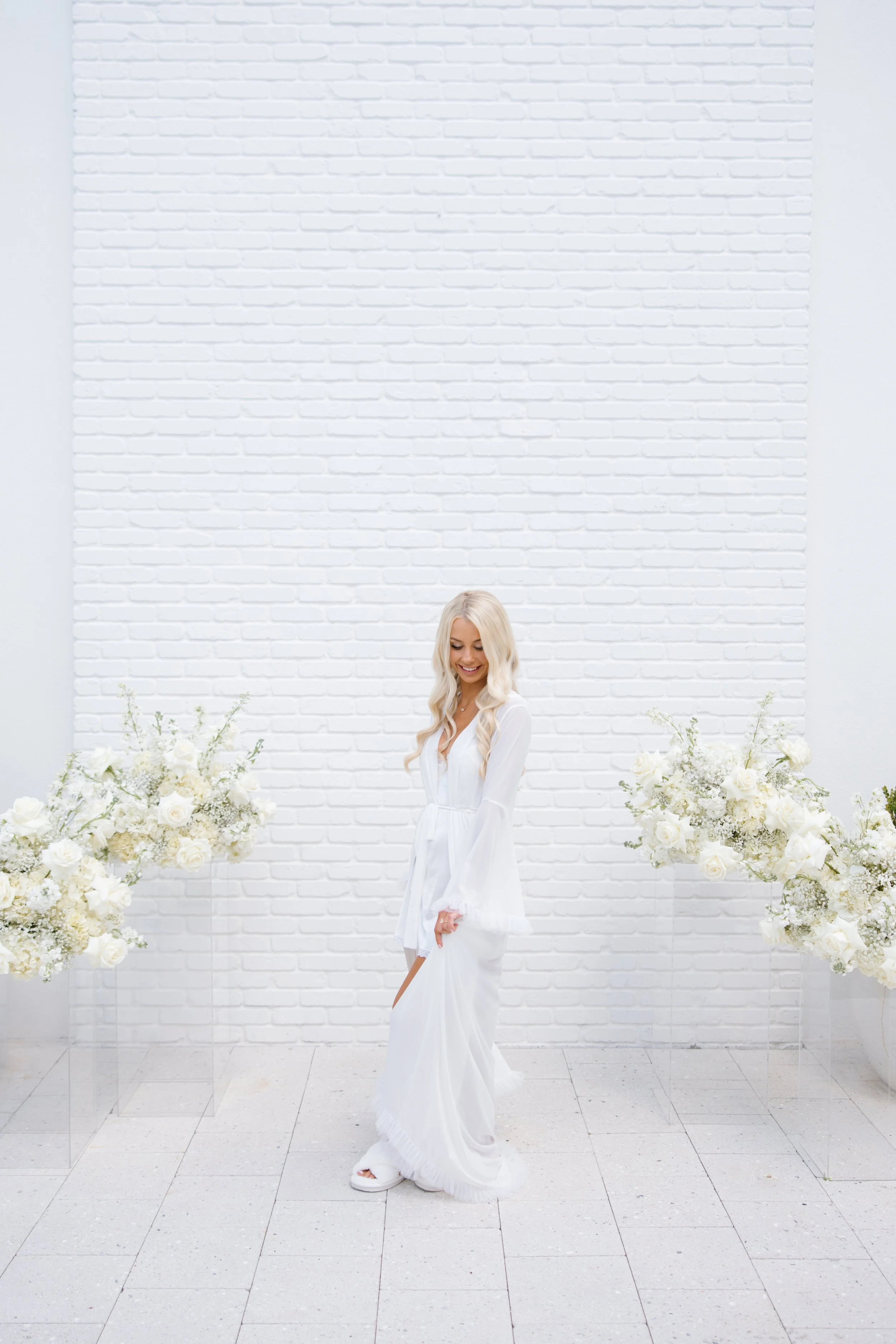 A woman with long, wavy blonde hair, smiling and wearing a white dress, stands among large white flower arrangements against a white brick wall.