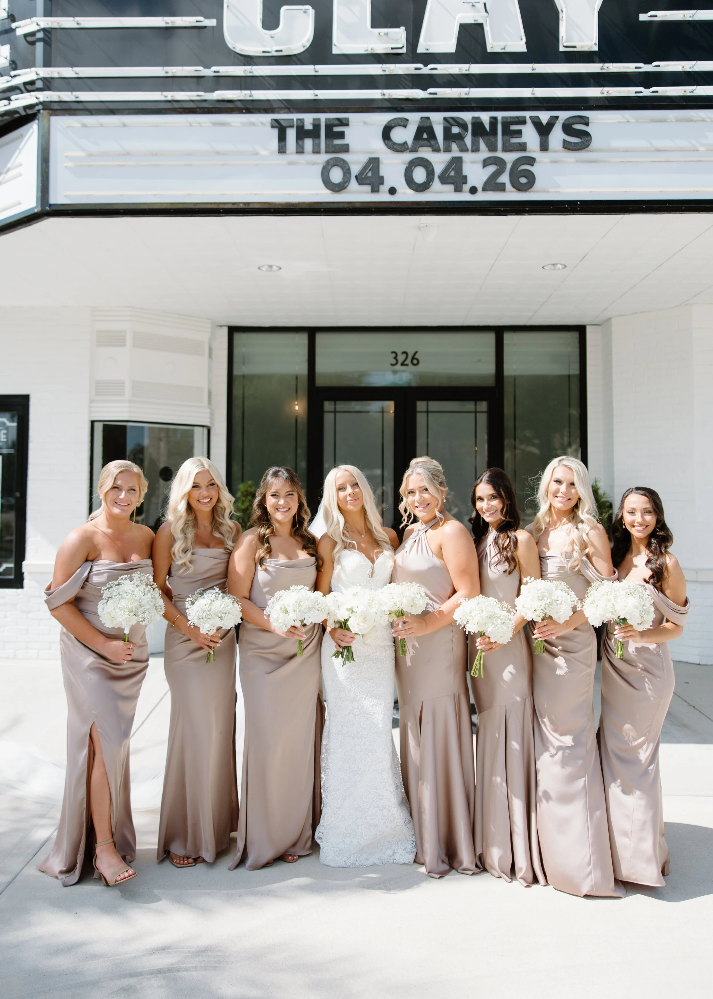 A group of women in bridesmaid dresses and a bride in a white wedding gown posing outdoors in front of a building with a marquee sign that reads "The Careys" and a date of April 26.