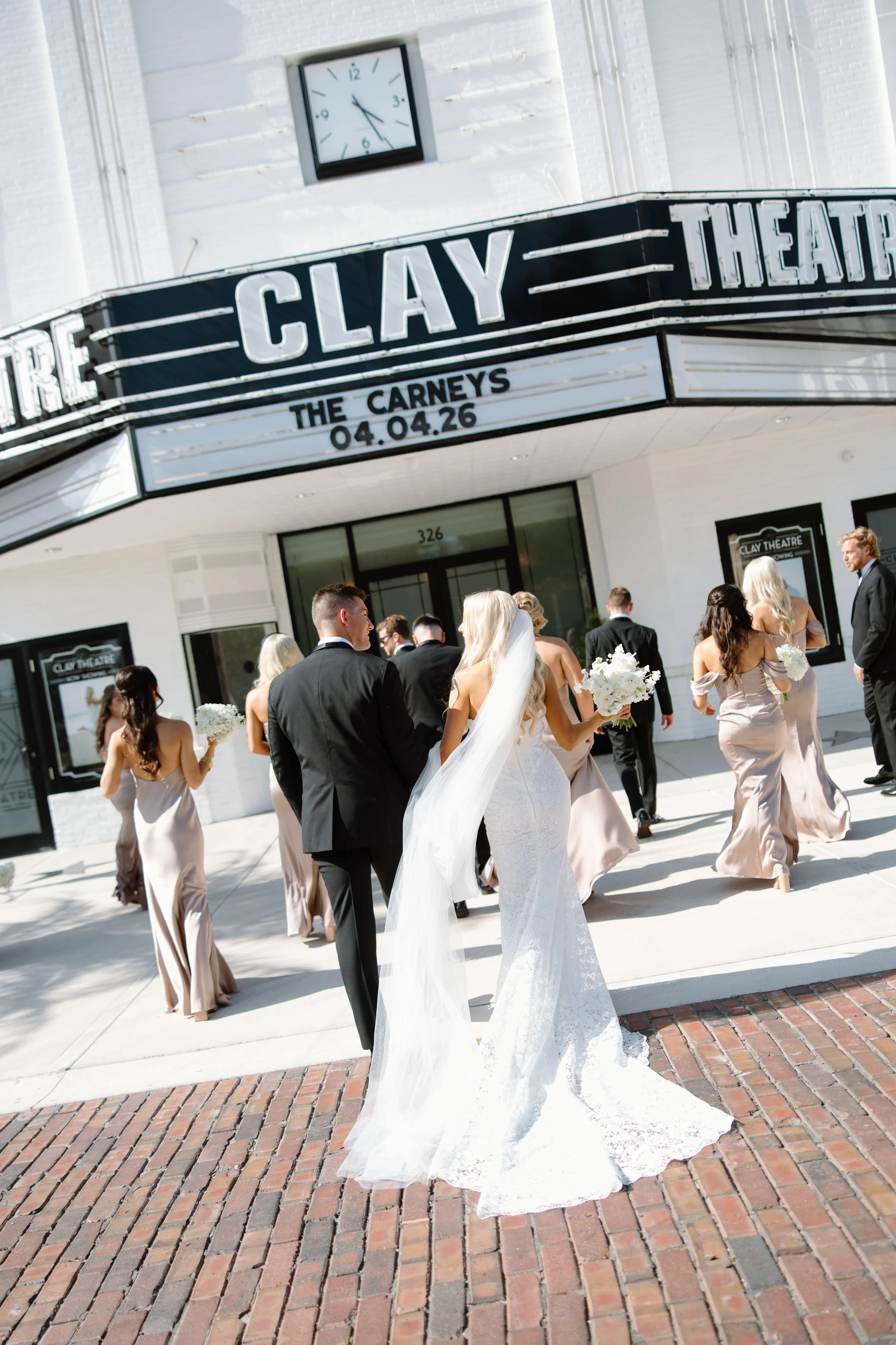 A group of people in formal attire, including a bride in a white gown with a long train and veil, is walking outside a theater building. The theater marquee displays the name 'Clay' and the event date '04.04.26'. The background features a white brick