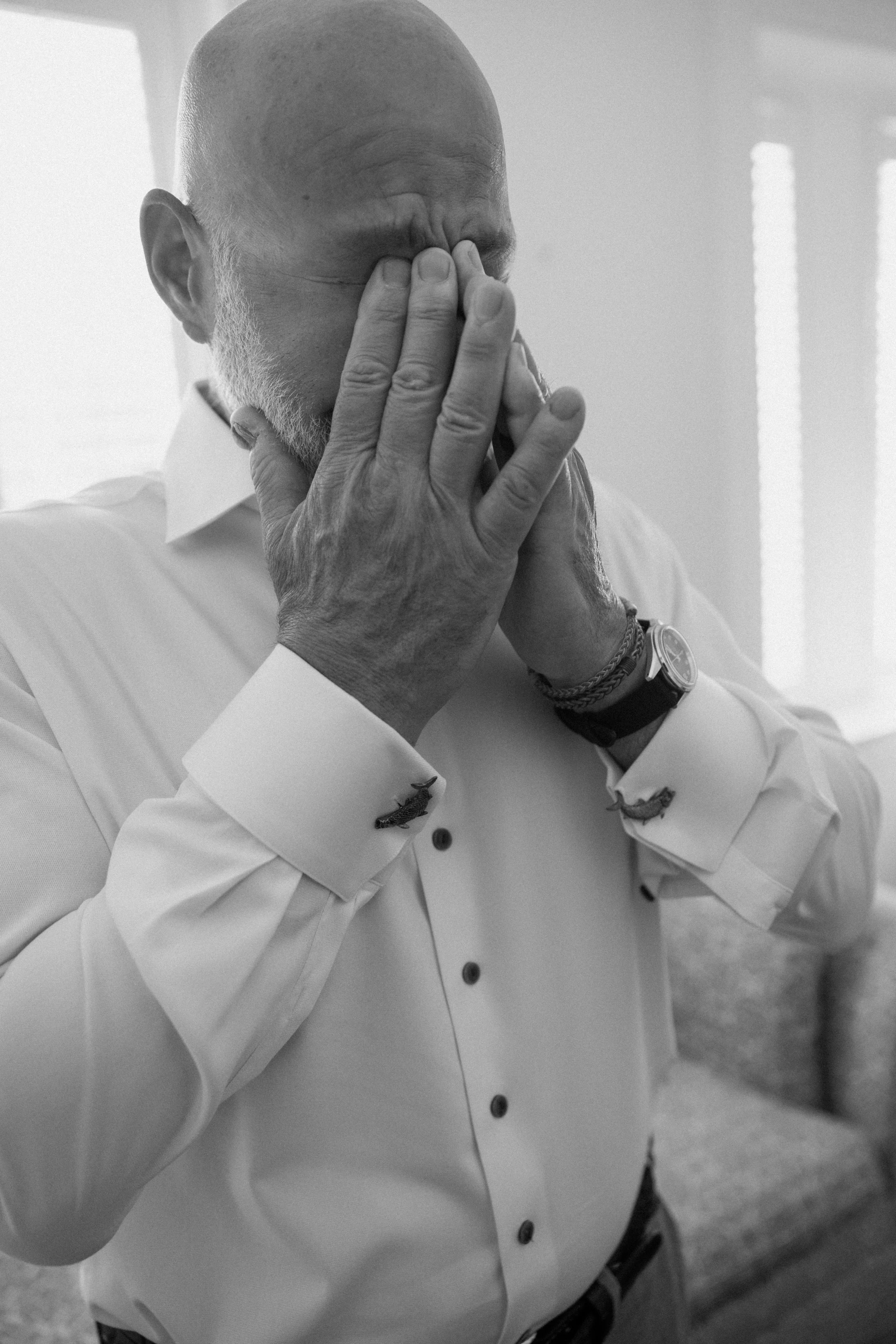 Black and white photo of a bald man with a beard, wearing a white shirt with rolled-up sleeves and a wristwatch and bracelets, holding his face with both hands, covering his eyes.