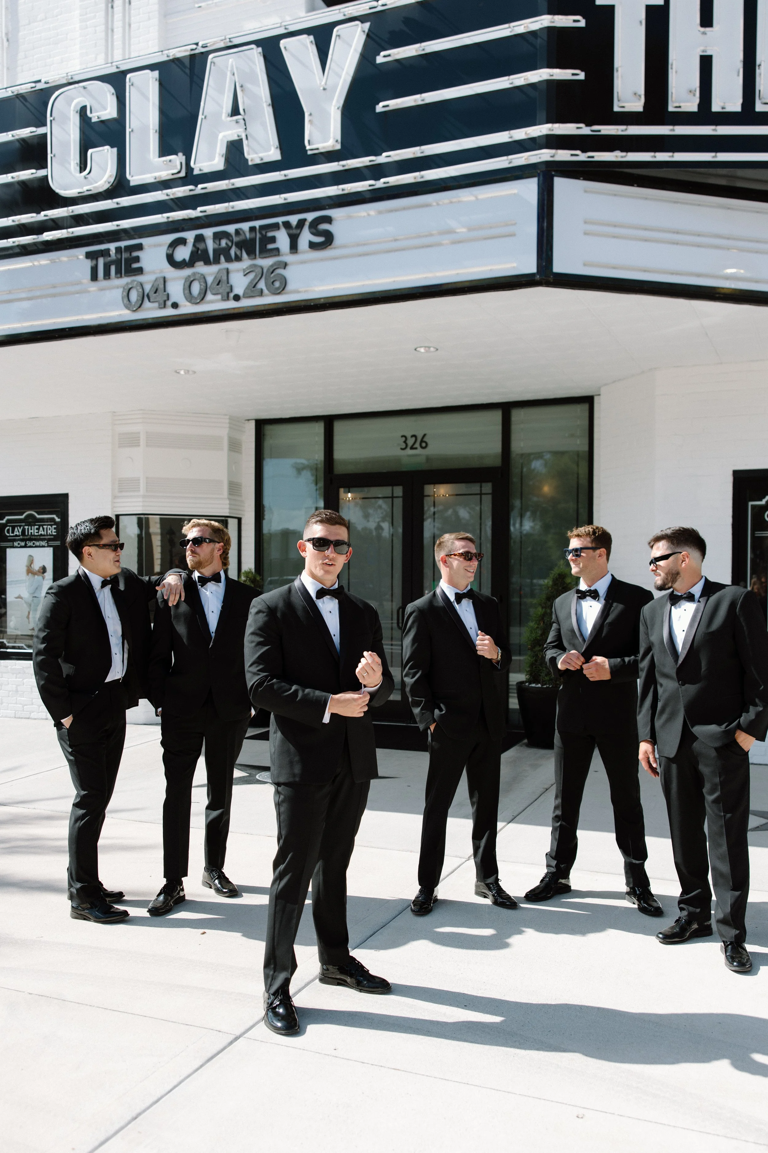 Group of six men in tuxedos and sunglasses standing outside a theater with a marquee displaying the name and date of a show.