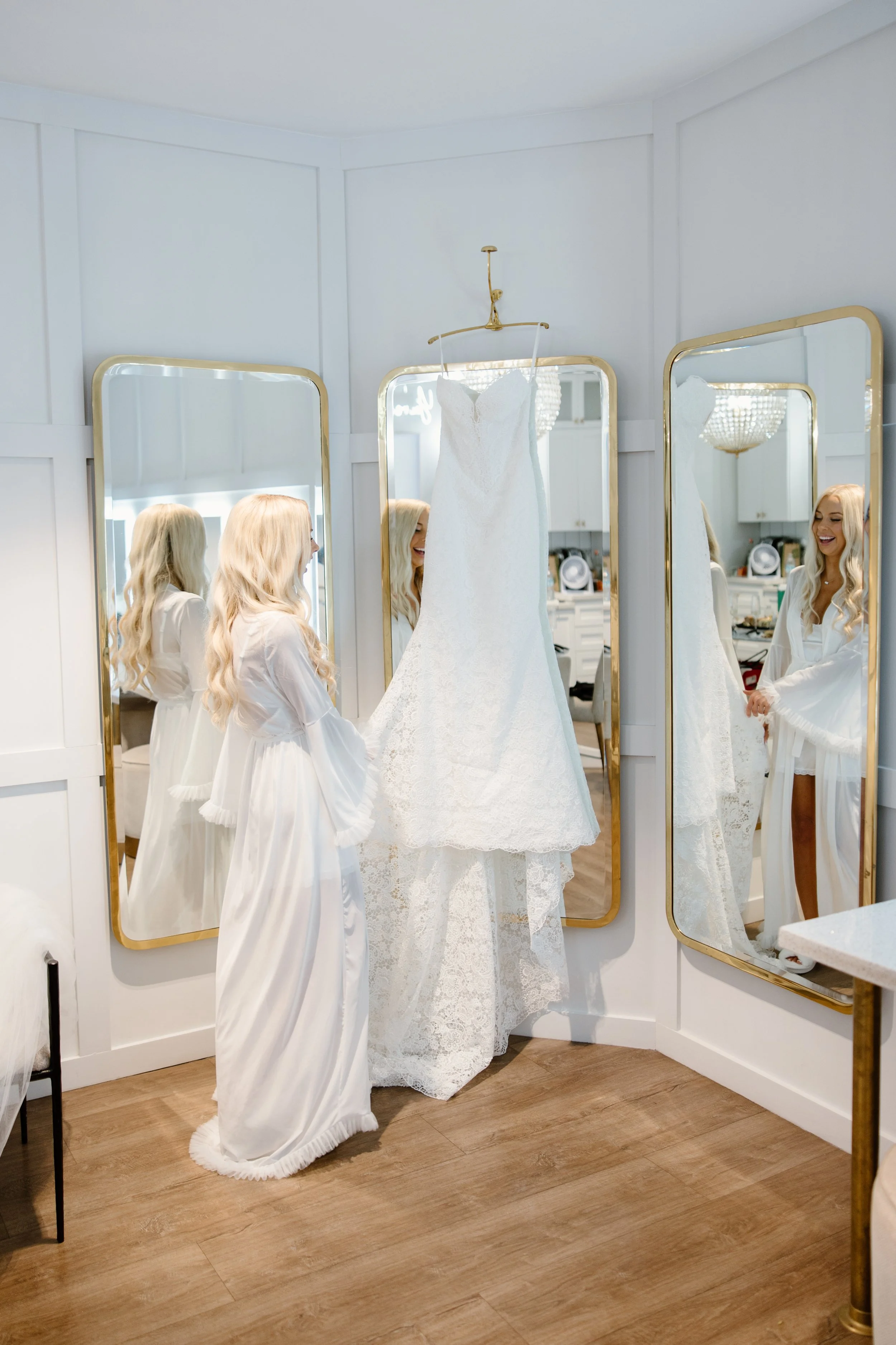 A woman with long blonde hair in a white robe is smiling while looking at her reflection in a mirror. A white wedding dress hangs on a hook above her, and she is holding the dress's train. The scene appears to be in a bright, modern room with wooden 