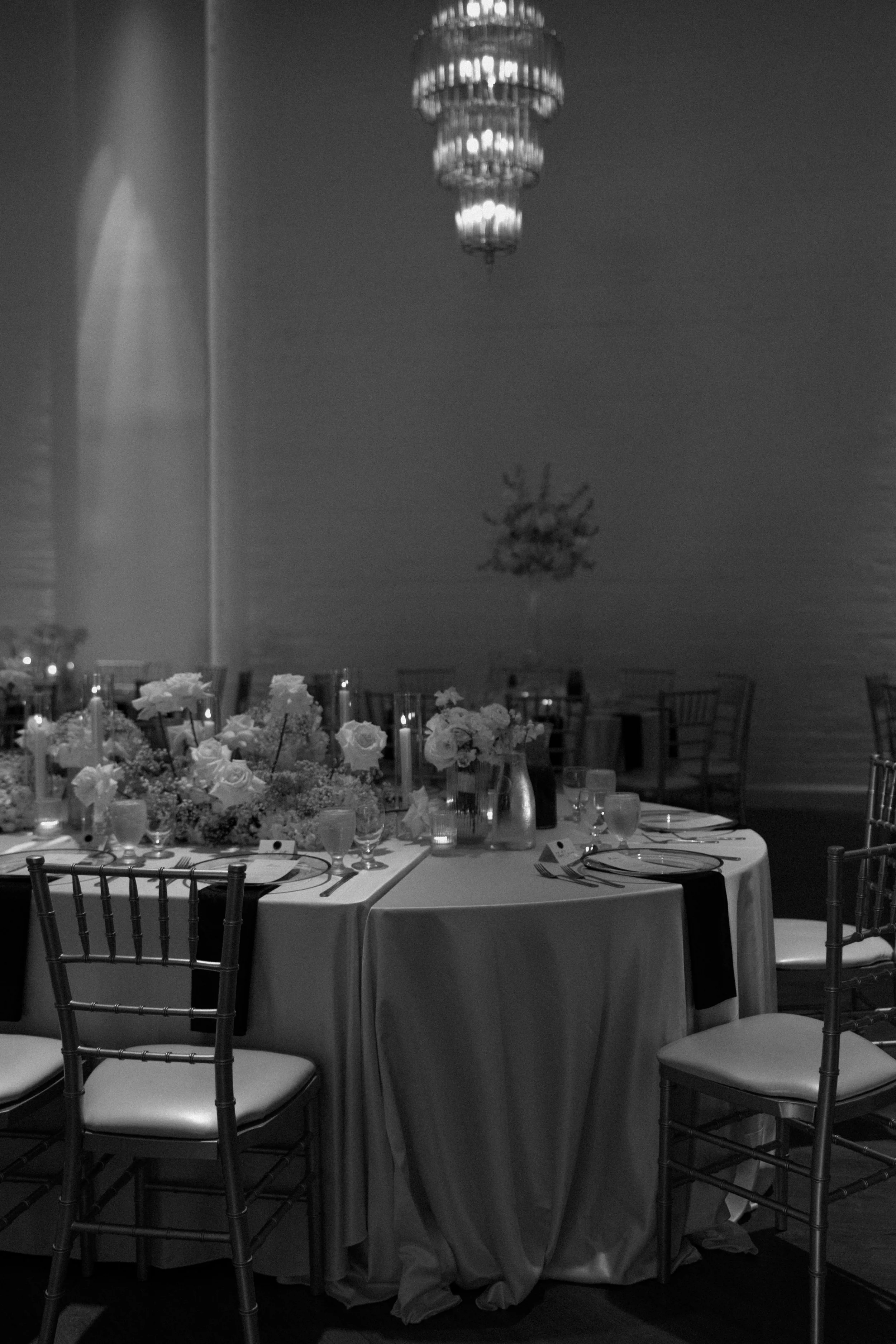 Elegant indoor dining setup with round table covered in a white tablecloth, floral centerpiece, candles, and place settings. Surrounding chairs are dark wood with white cushions. The background features a chandelier hanging from the ceiling and subdu
