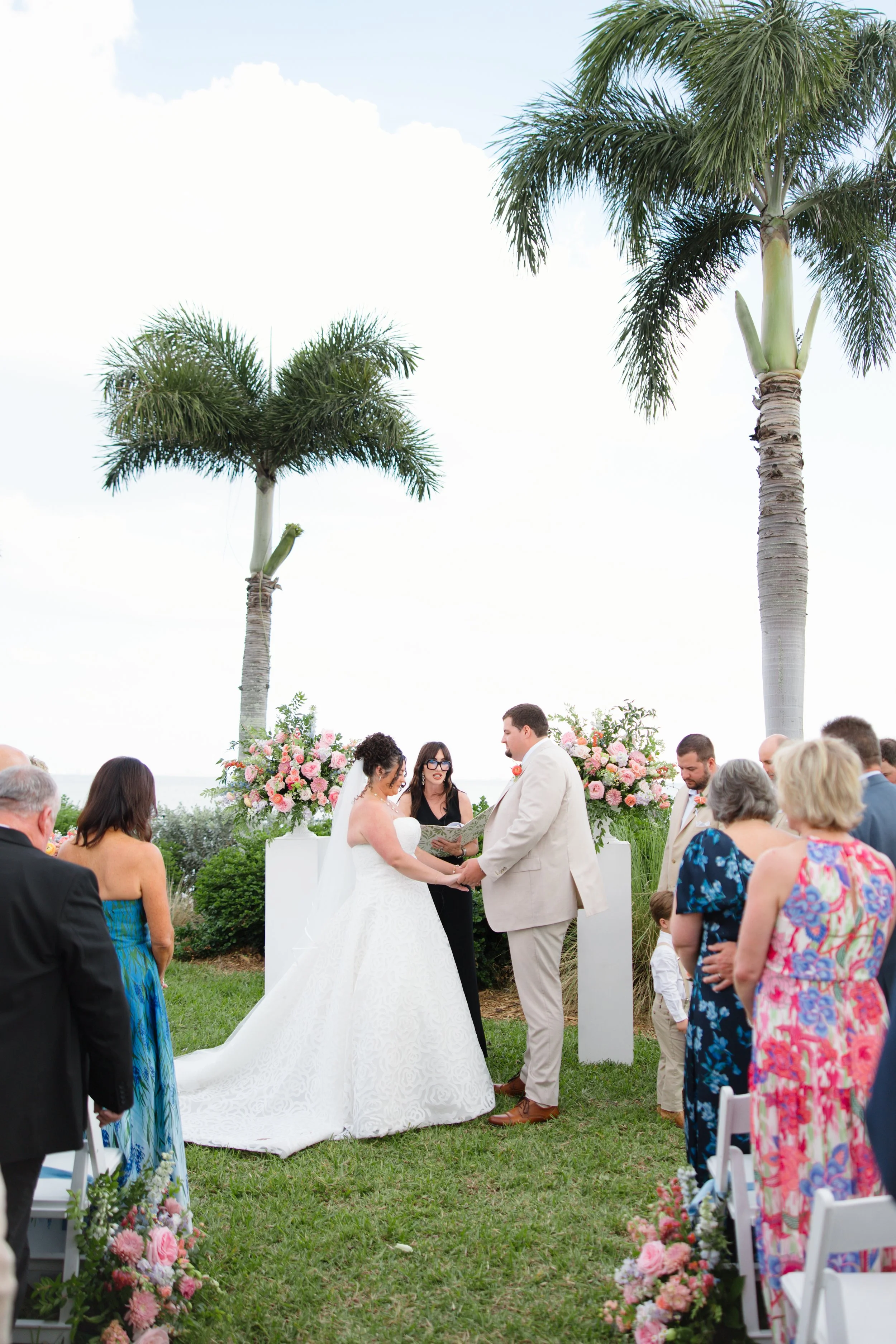 A wedding ceremony outdoors with the bride and groom holding hands and exchanging vows, surrounded by floral arrangements and guests, under tall palm trees.