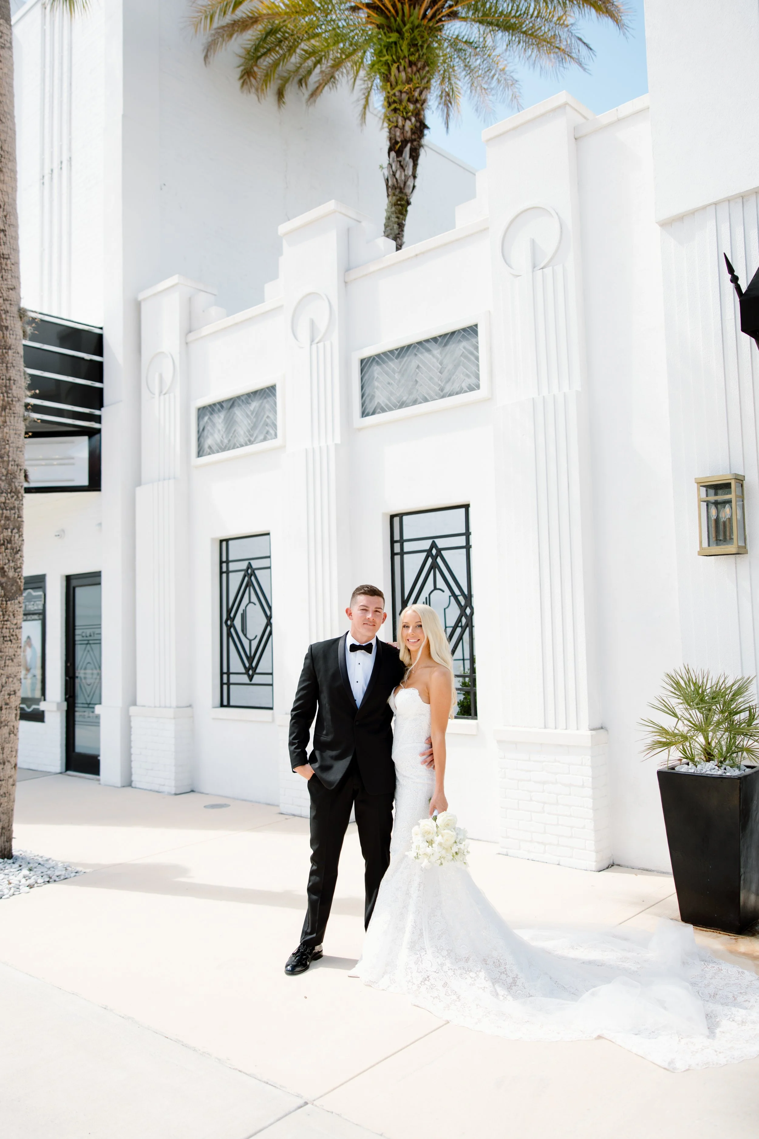 A newlywed couple stands outside a white building with art deco design elements. The groom is dressed in a black tuxedo with a bow tie, and the bride is in a strapless white wedding gown holding a bouquet of white flowers. A tall palm tree is visible