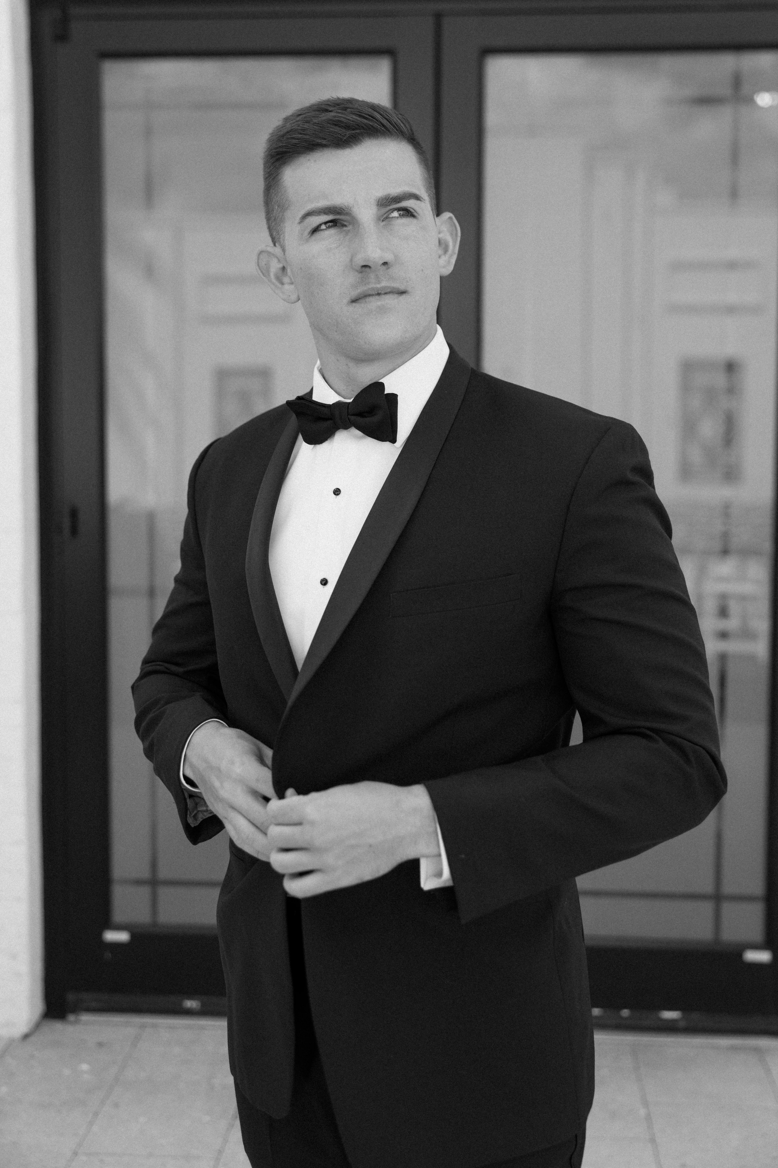 A young man in a tuxedo with a bow tie standing indoors with a serious expression, looking to his left.
