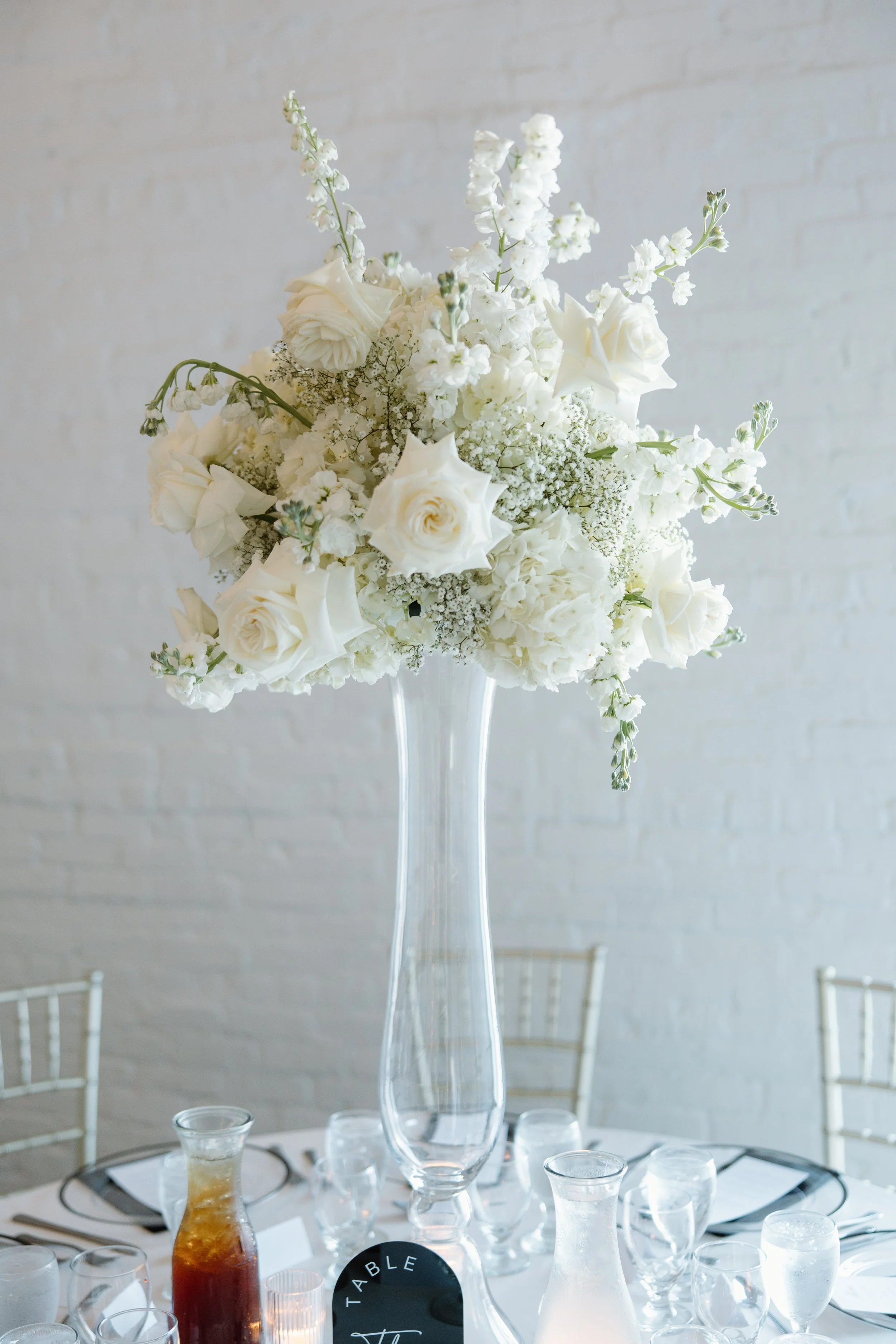 A tall glass vase with a bouquet of white flowers, including roses and various small white blossoms, on a table decorated for an event.