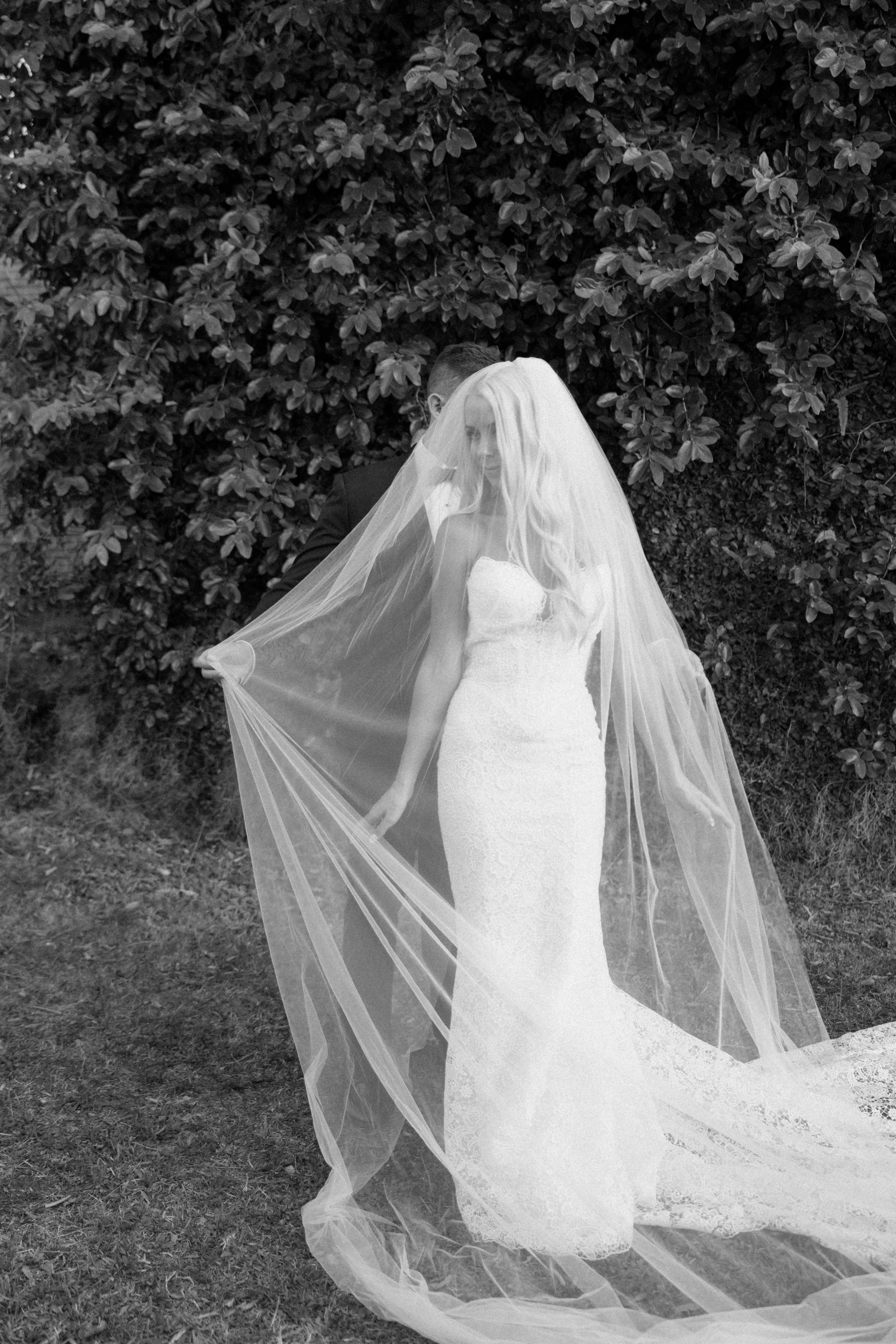 A bride wearing a strapless lace wedding gown and long veil standing outdoors in front of dense bushes.