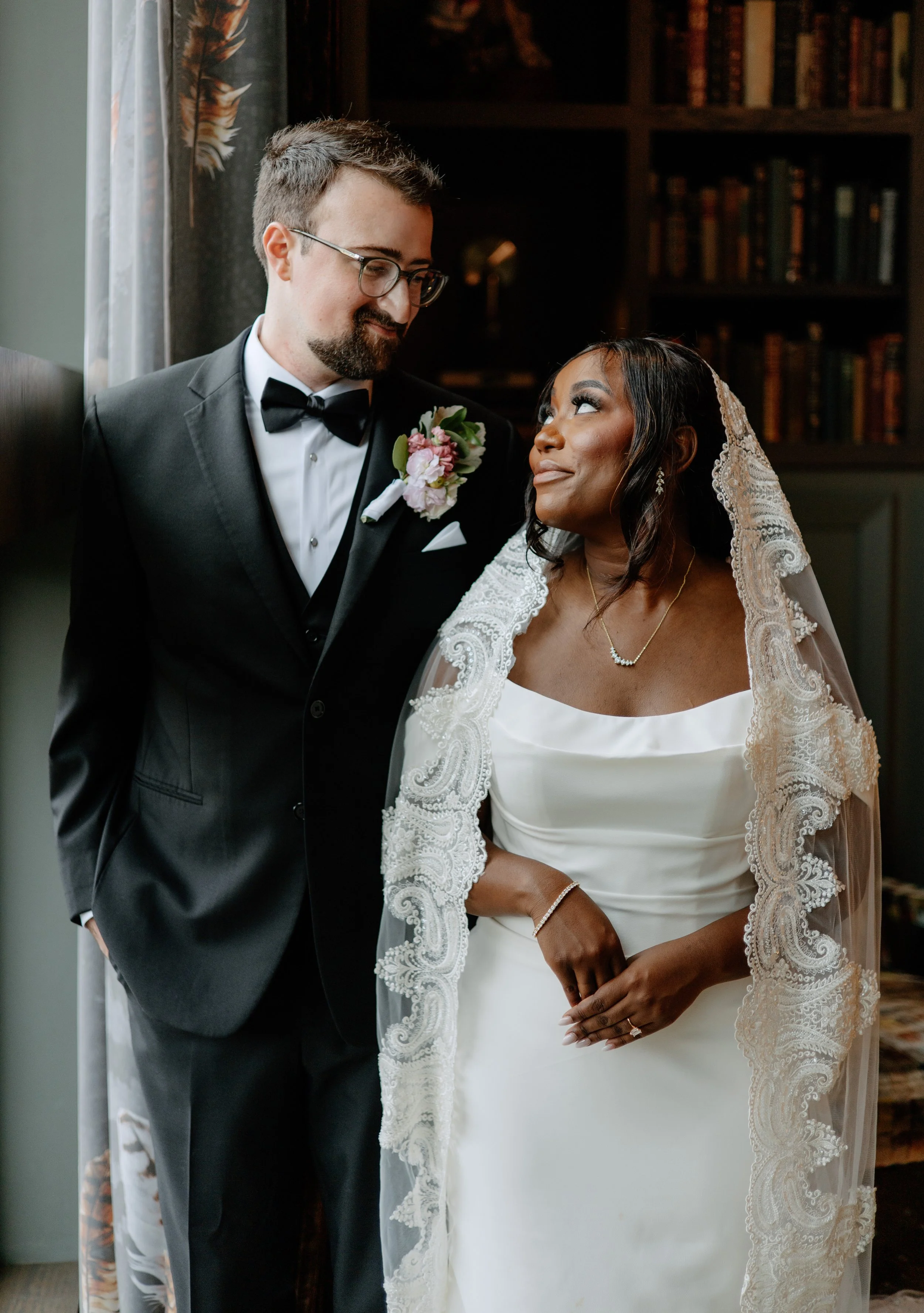 A bride and groom standing close to each other in a room with dark shelving and curtains. The groom is dressed in a black tuxedo with a bow tie and a floral boutonniere. The bride wears a white gown with lace details and a lace veil, and is looking up at the groom with a smile.