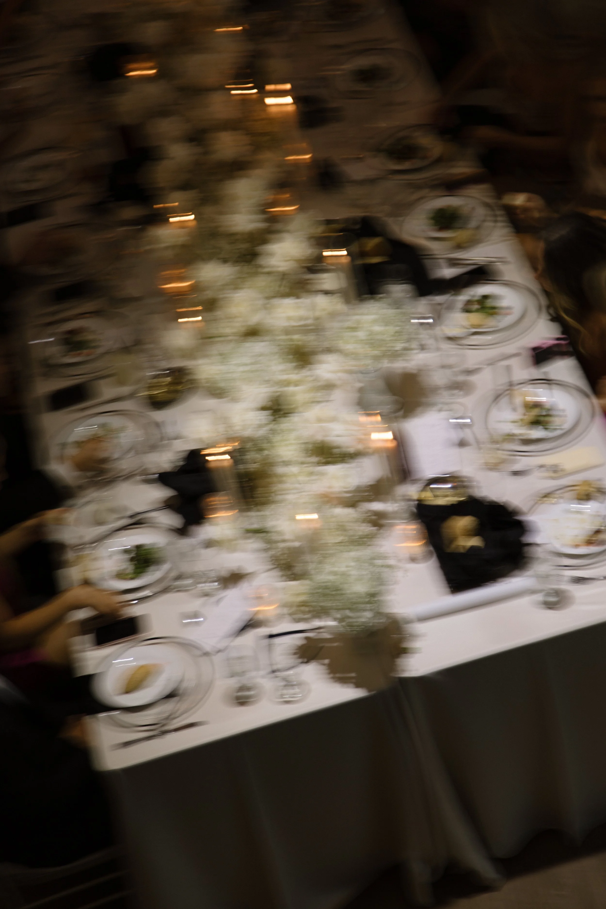 A blurry image of a banquet table set with plates, glasses, utensils, and floral centerpieces, with people seated around.