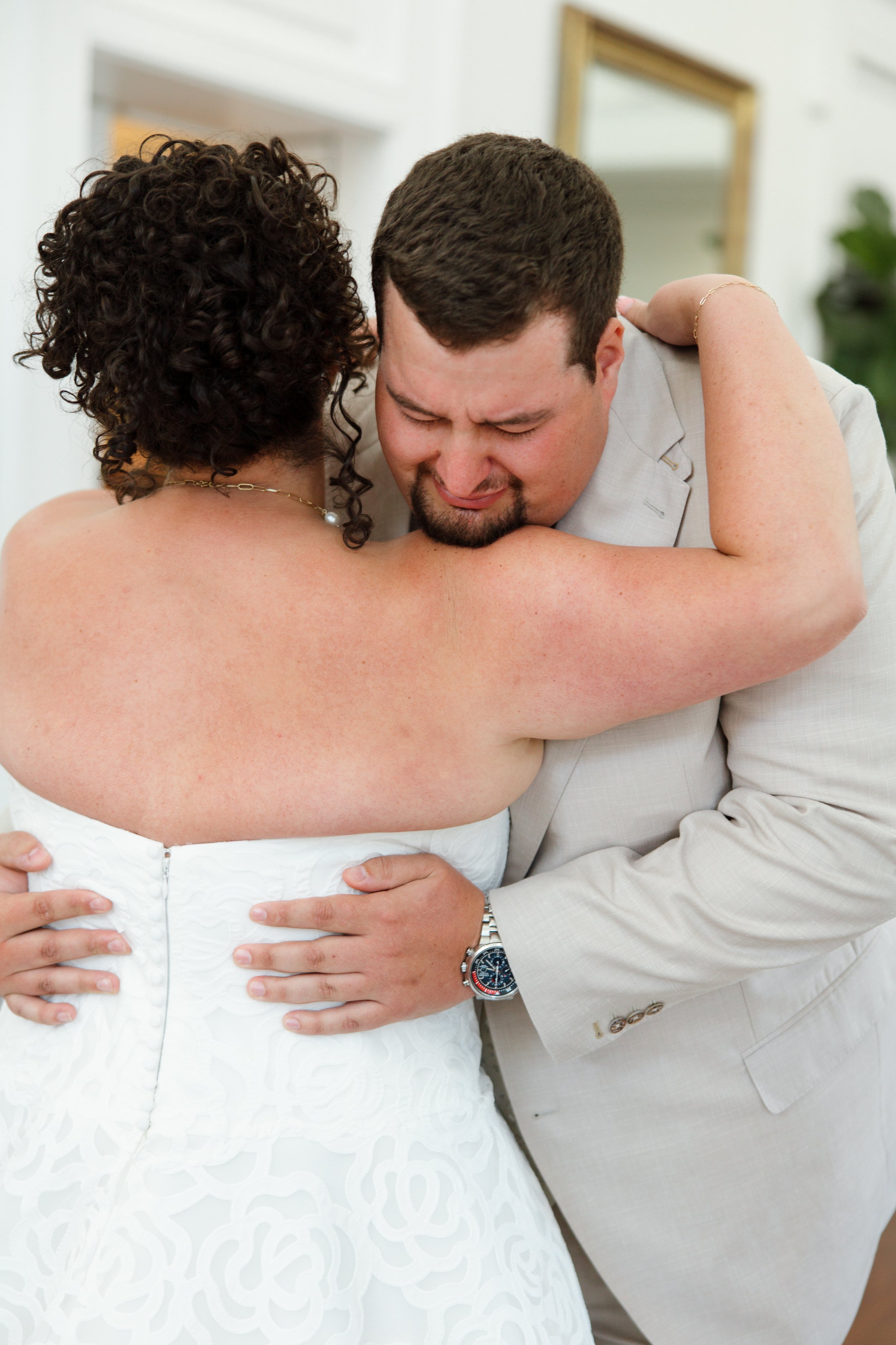 A couple is embracing, the woman in a strapless white dress and the man in a light gray suit. They are holding each other close, with heads touching and eyes closed.