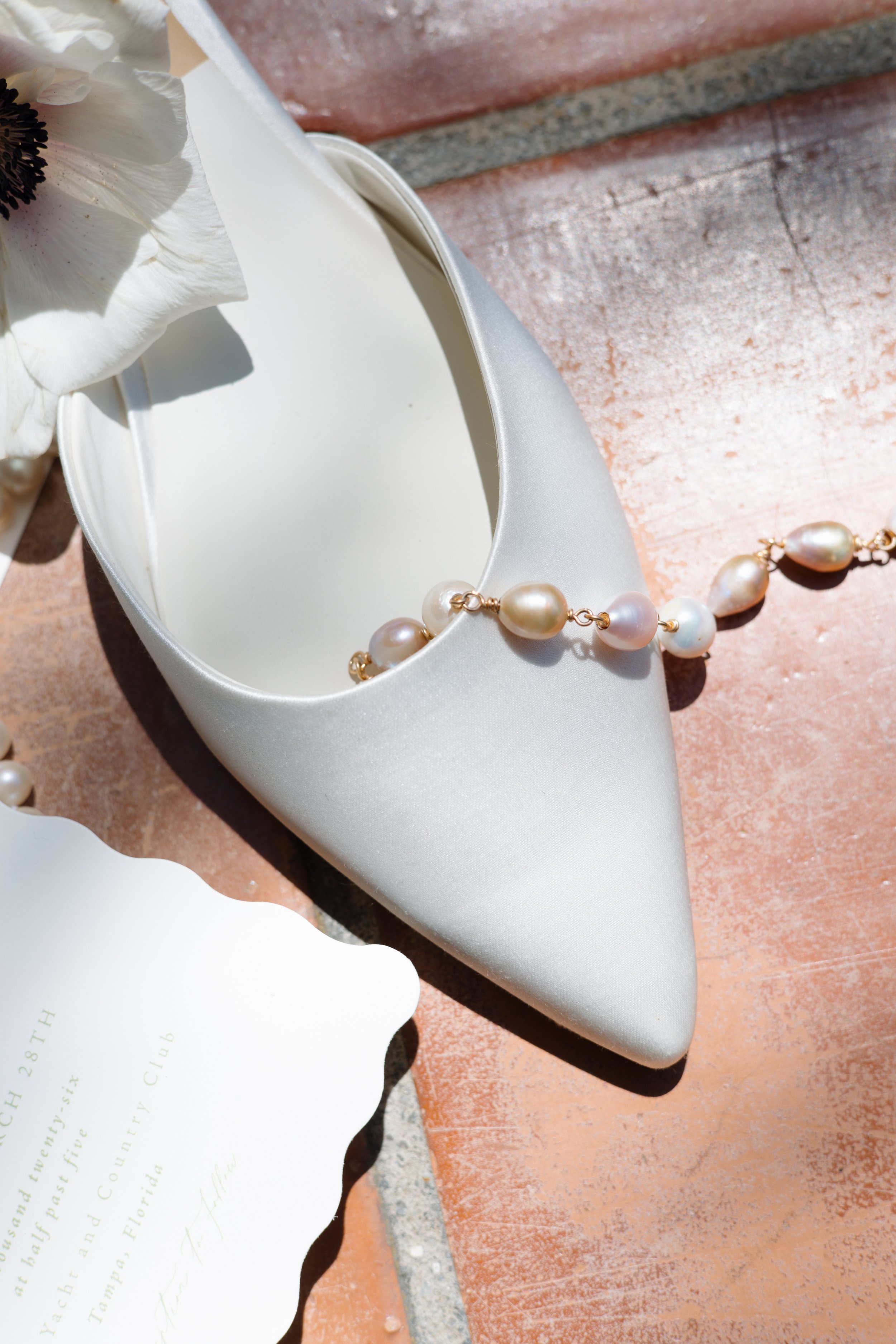 Close-up of a white satin high-heeled shoe with a pearl necklace draped over it, on an orange-red brick surface.
