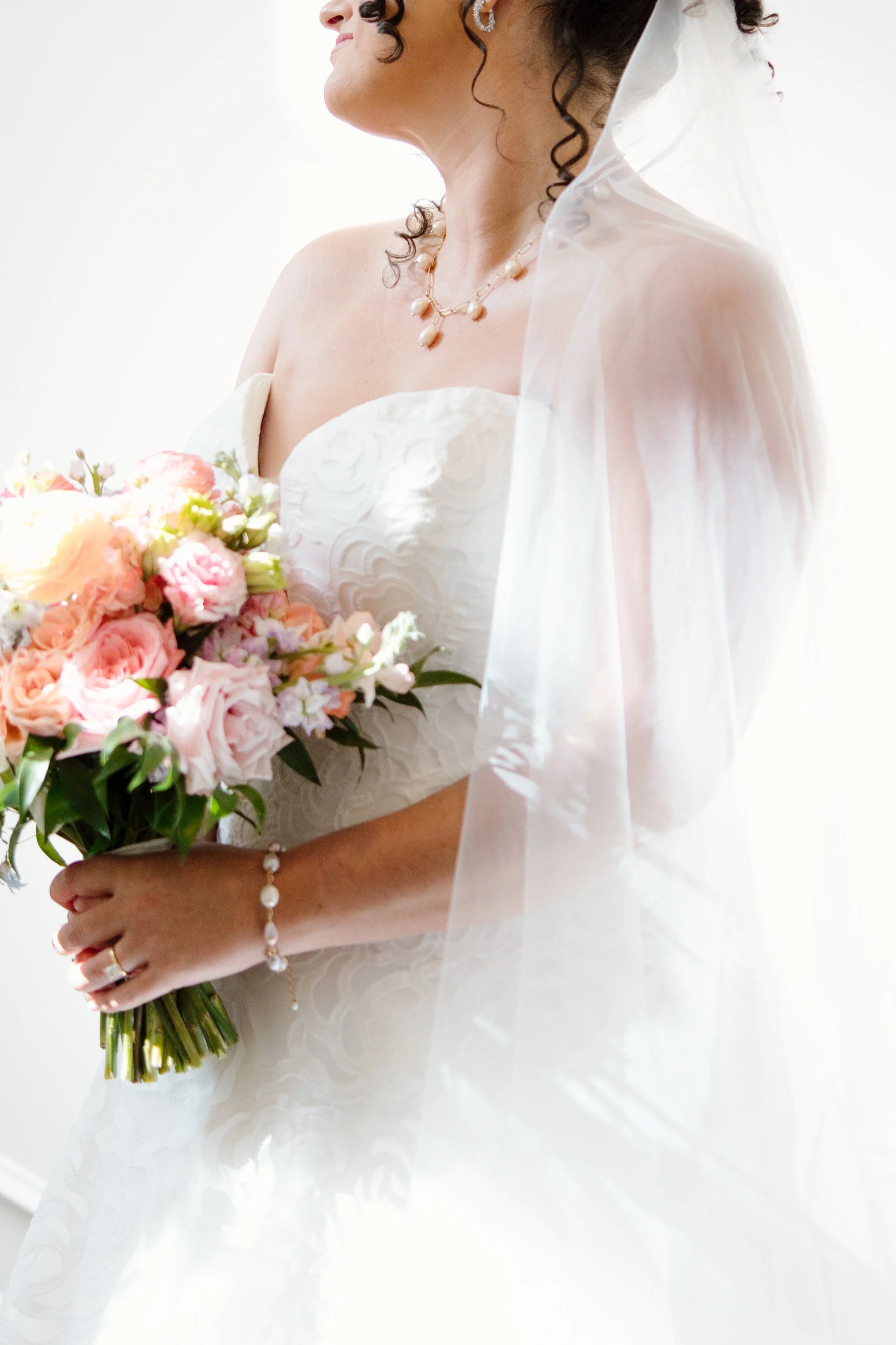 A bride holding a bouquet of pink and peach flowers, wearing a strapless white wedding dress with lace details, jewelry including a necklace, earrings, a bracelet, and rings, with a veil covering her head and shoulders, against a white background.