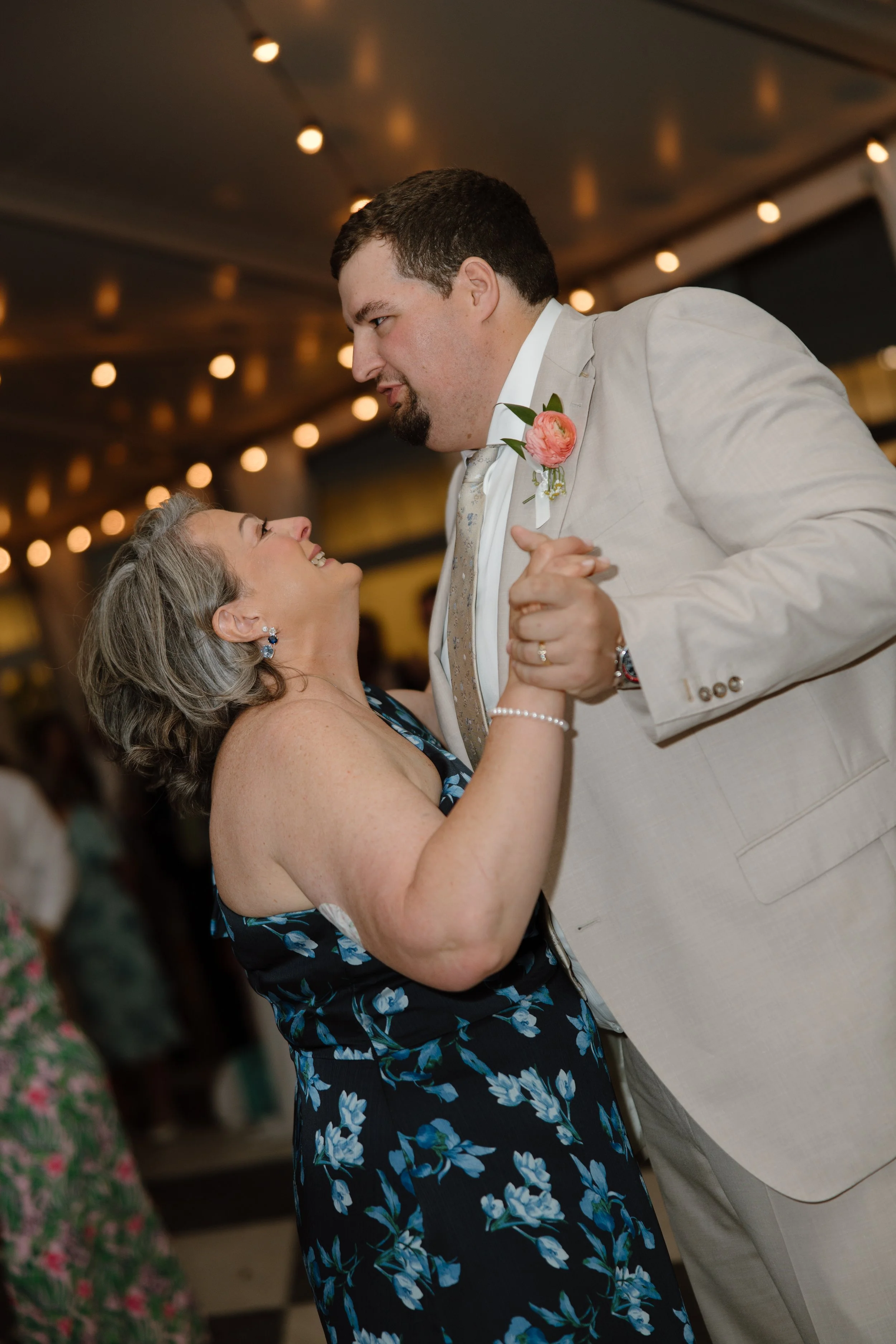 A woman and a man are dancing together at a wedding reception, smiling up at each other. The woman is wearing a floral dress and the man is in a beige suit with a pink flower boutonniere. They are holding hands and looking into each other's eyes, wit