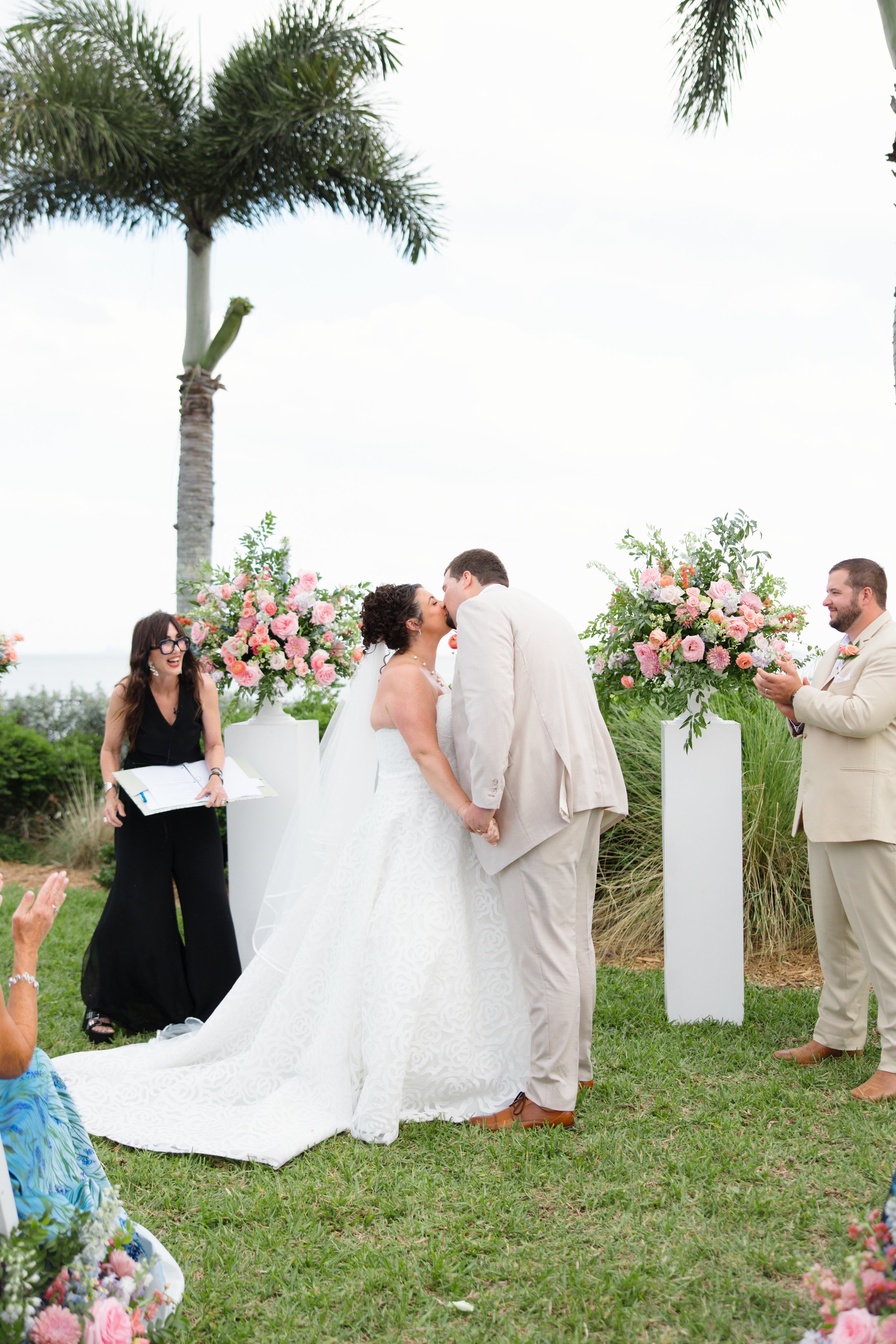 A couple kissing at outdoor wedding ceremony, surrounded by floral arrangements, officiant and guests, with palm trees and cloudy sky in background.