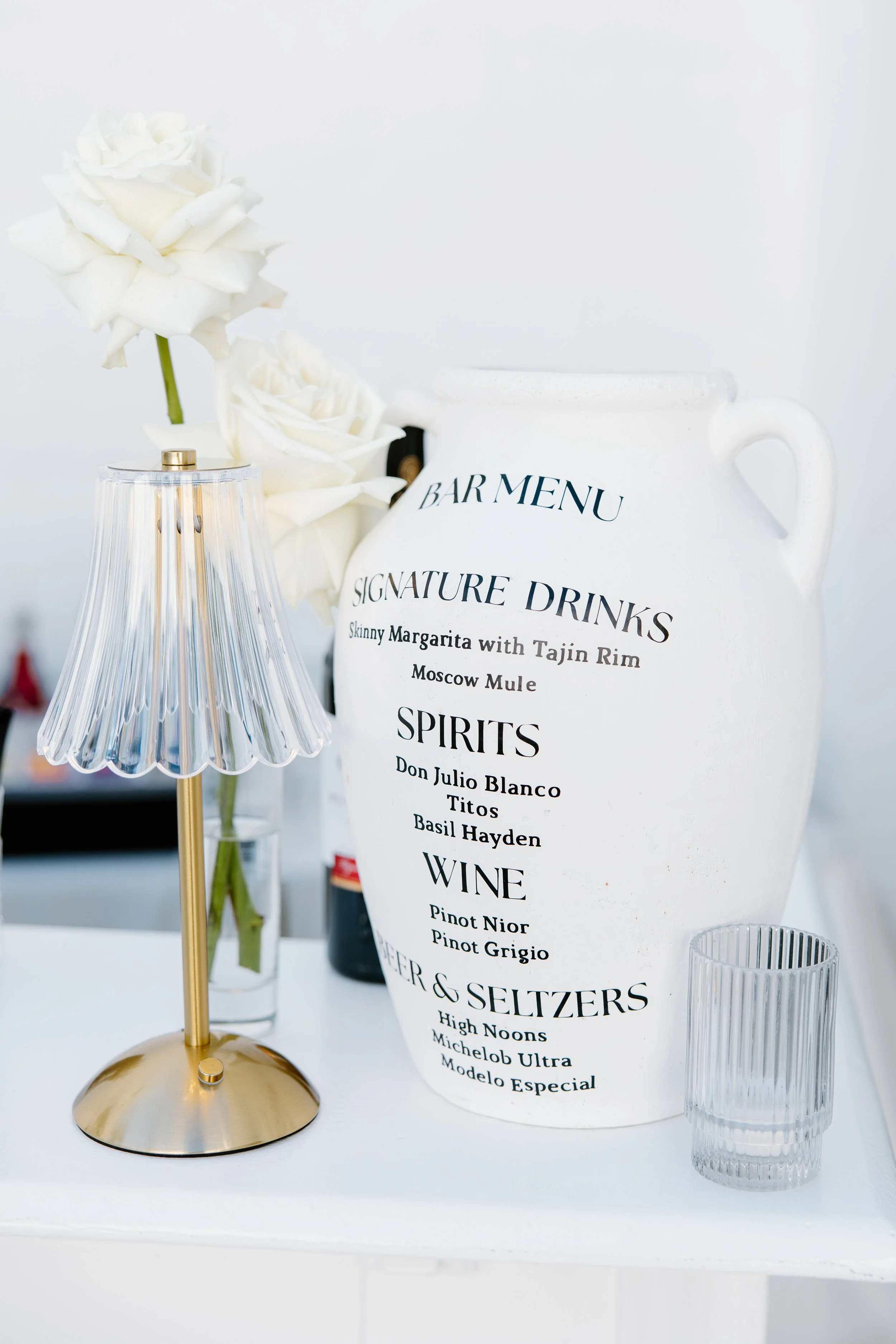 Decorative bar setup with a gold and glass table lamp, white roses in a vase, and a white ceramic pitcher with a printed menu listing drinks, spirits, and wines. Clear glass of water is also in the foreground.
