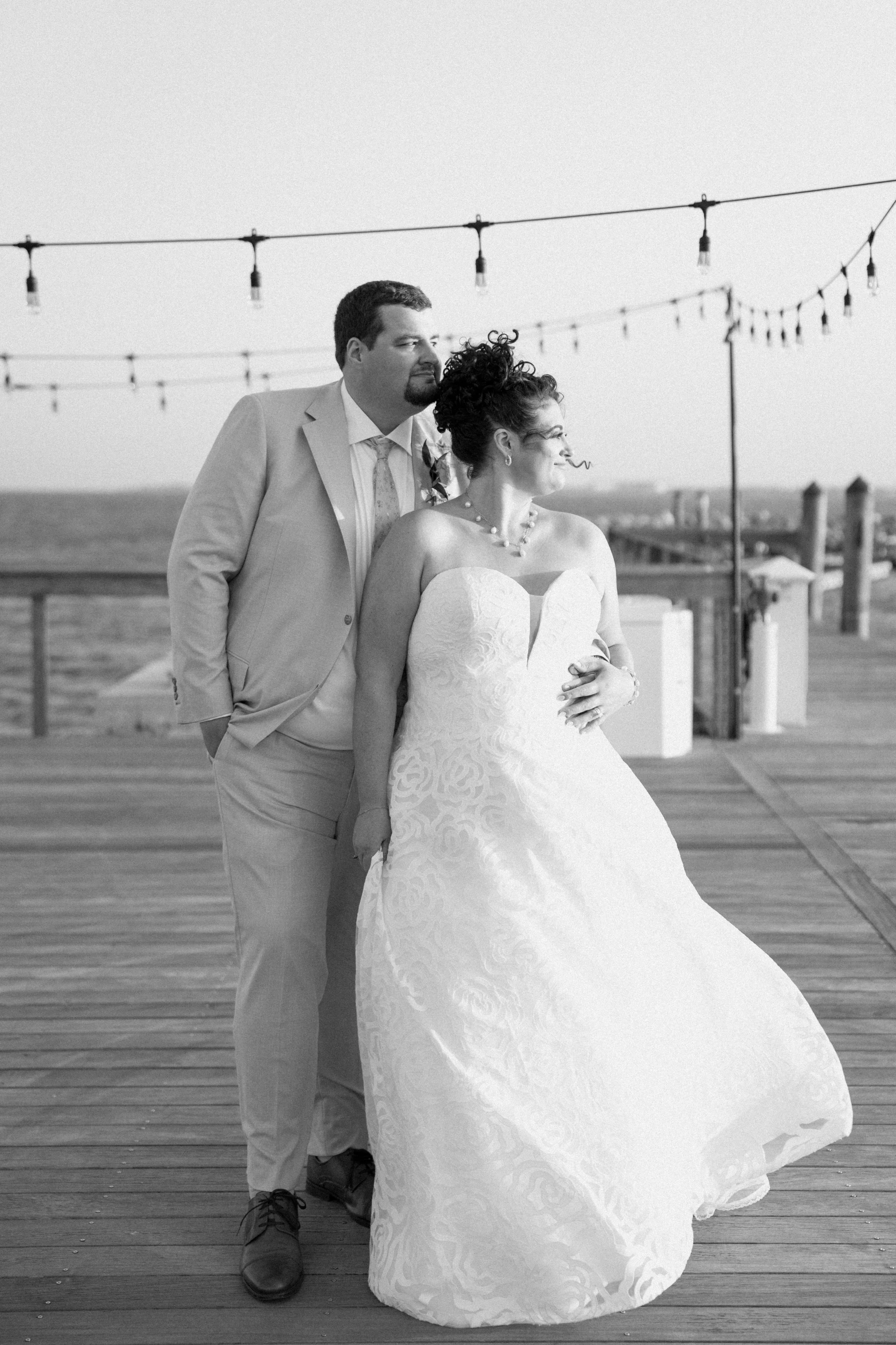 A black-and-white photo of a bride and groom standing on a wooden deck outdoors, with string lights overhead and a body of water in the background, during a wedding ceremony.