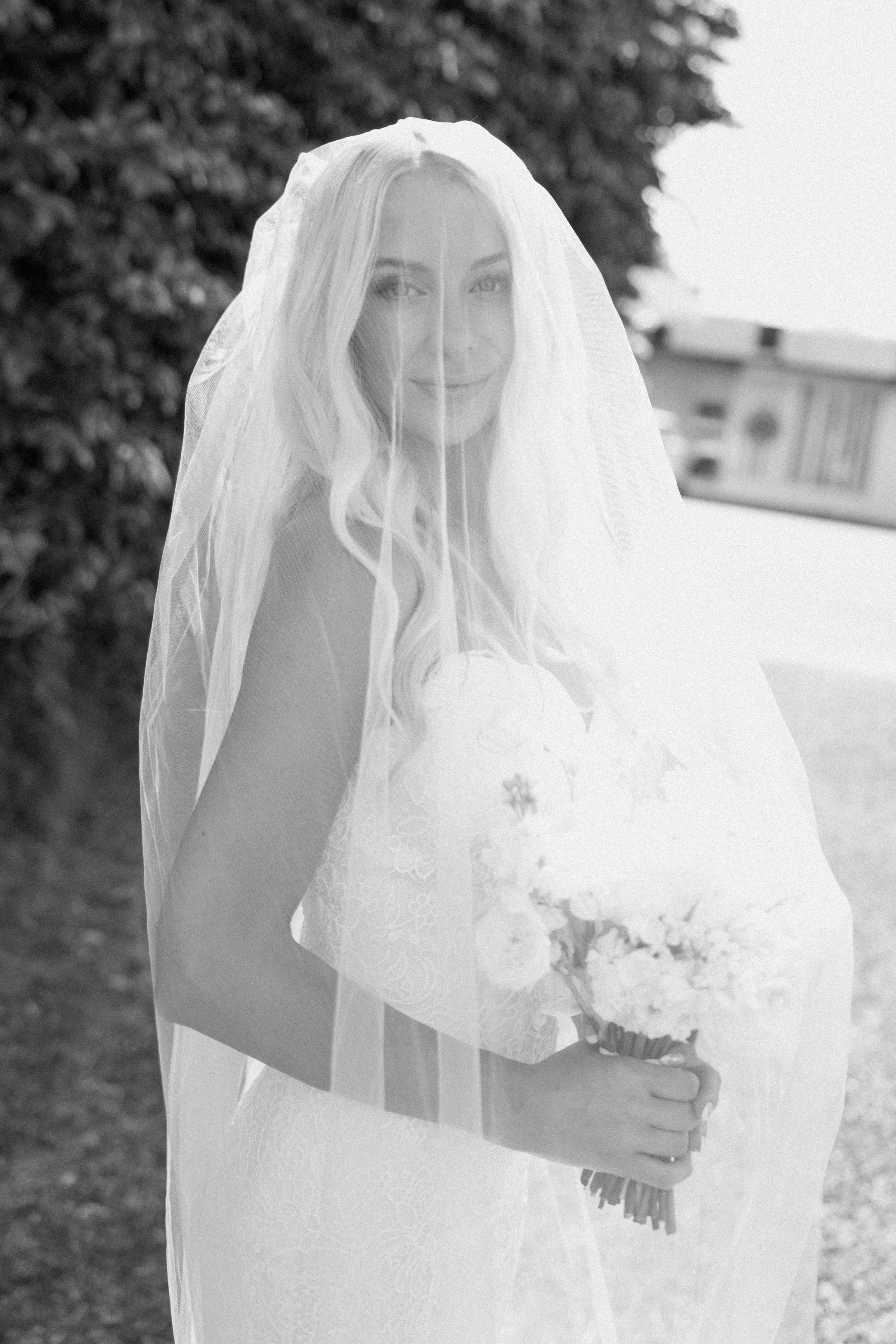 A woman in a wedding dress holding a bouquet, wearing a veil, standing outdoors with trees and a house in the background.