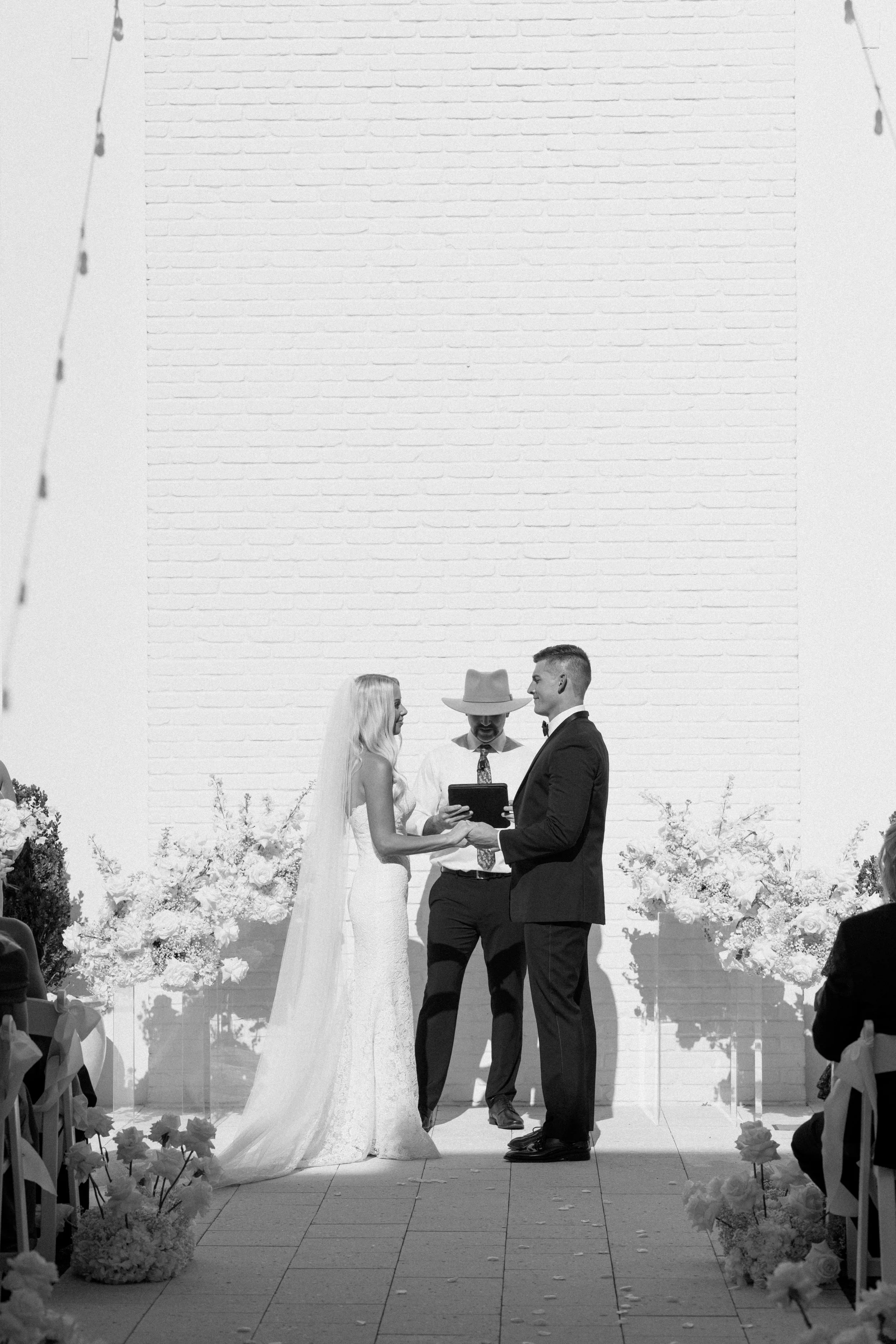 A black and white photo of a wedding ceremony with a bride, groom, officiant, and a large white brick wall background, floral decorations along the aisle.