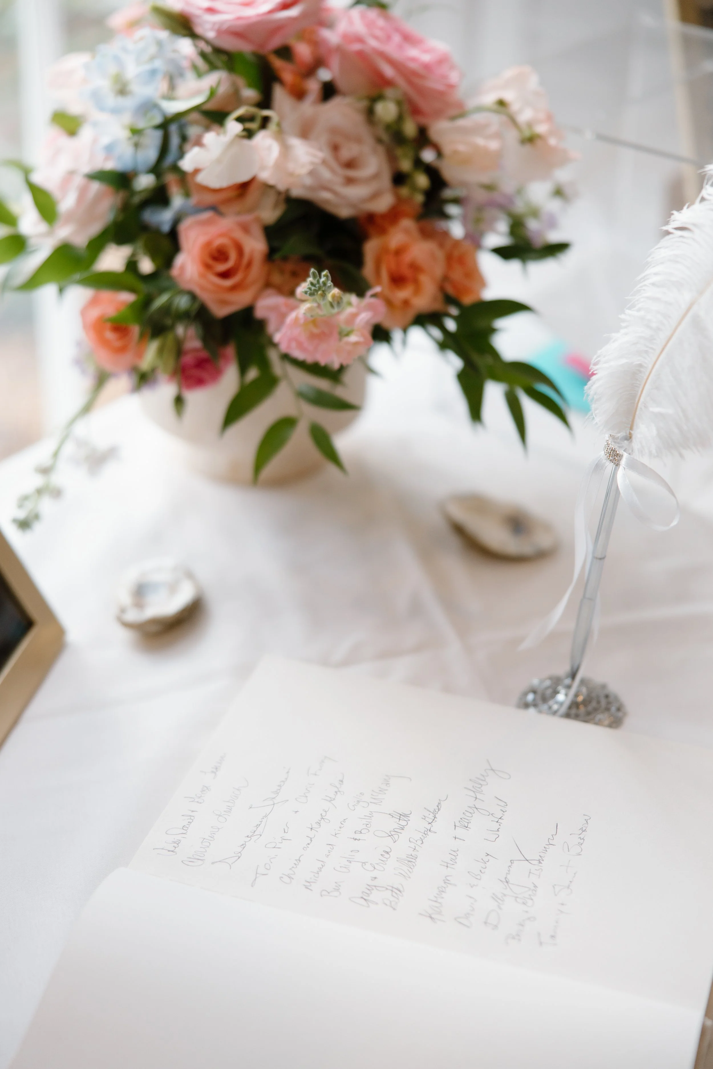 A blurred bouquet of pink, peach, and white flowers in a white vase on a table, with a handwritten letter or note on white paper in front, and a white feather decoration to the right.