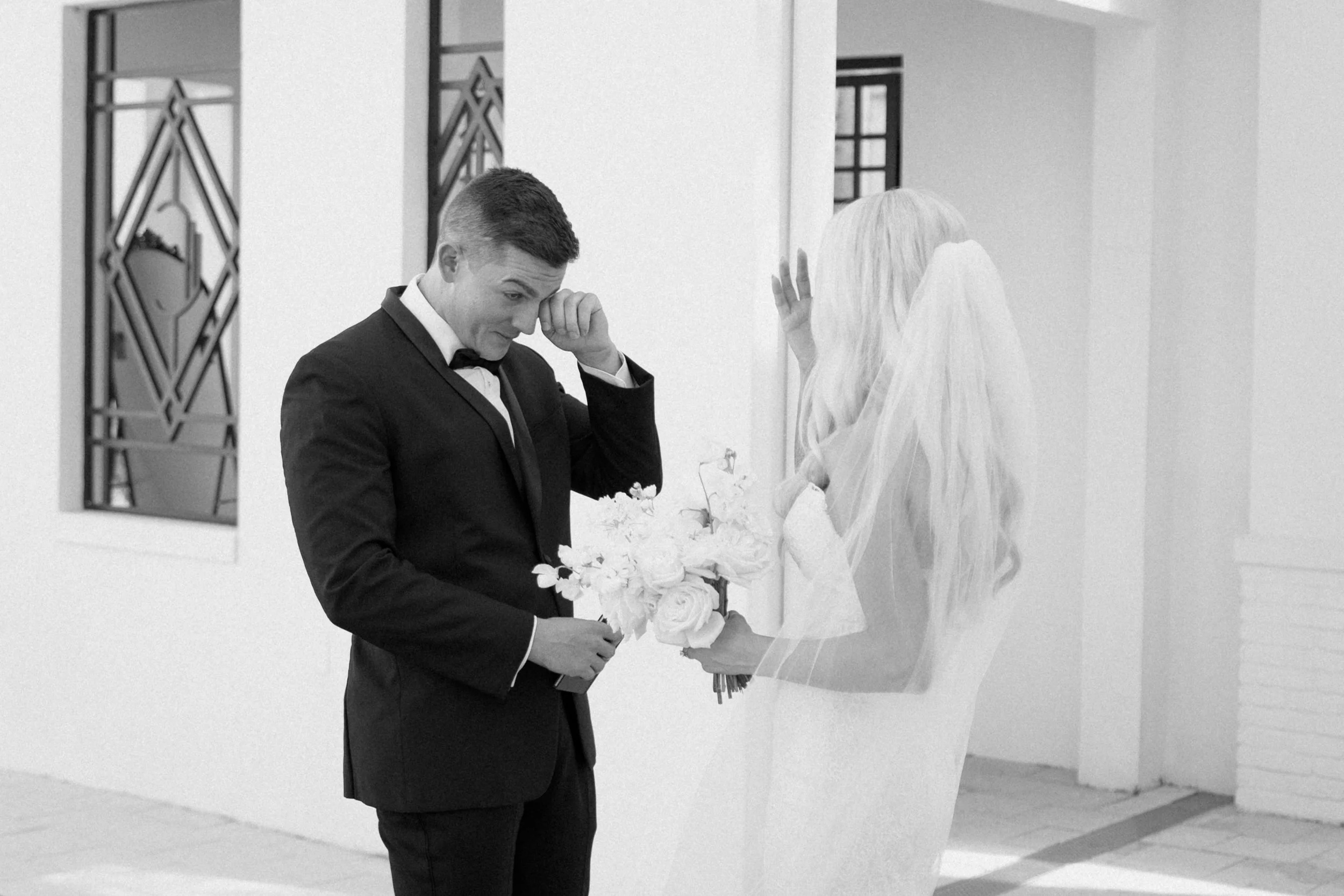 A man in a tuxedo holding a bouquet of white roses, standing in front of a woman dressed in a wedding gown, likely at a wedding or reception.