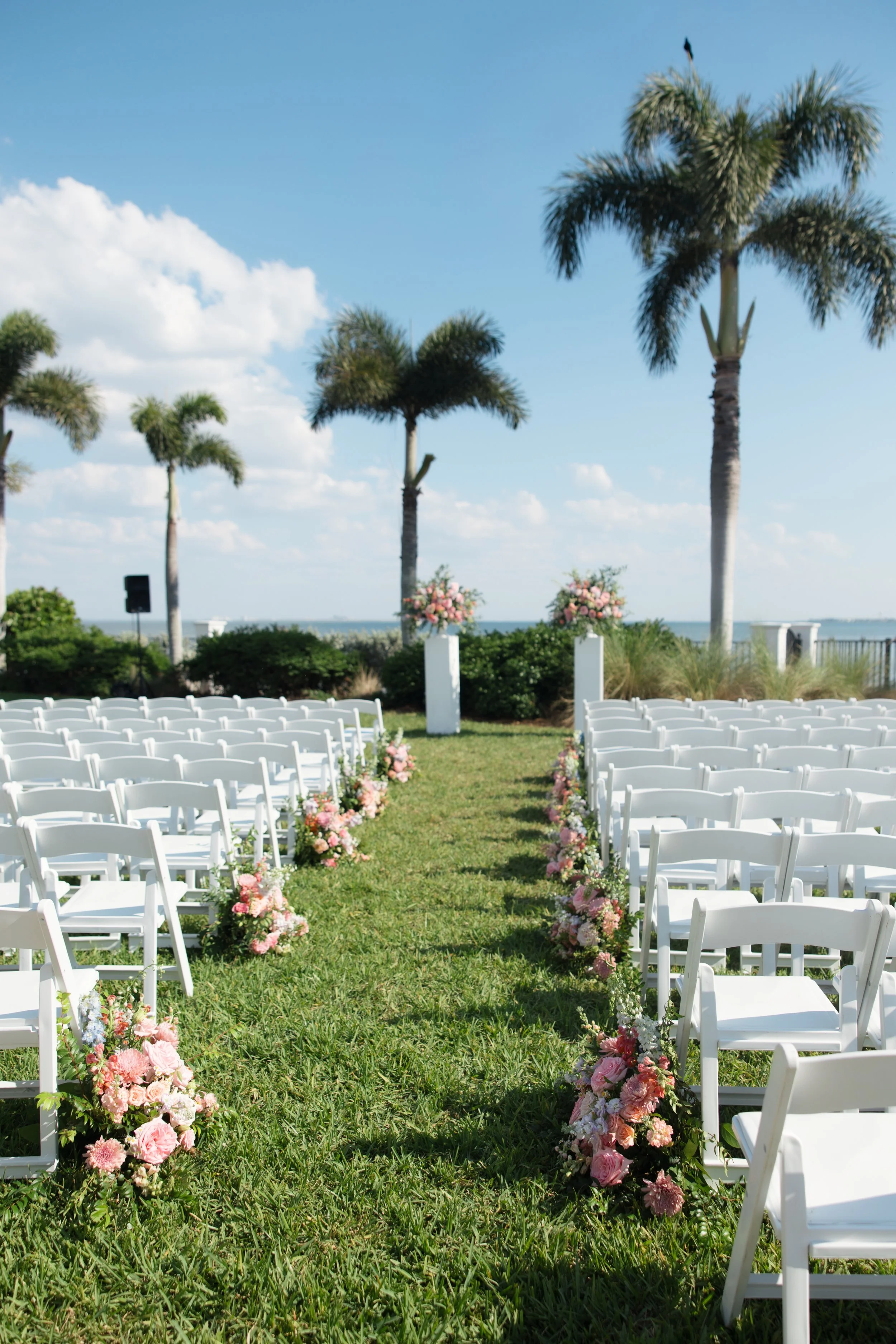 Outdoor wedding setup with rows of white chairs on green grass, decorated with pink and white flowers along the aisle, flanked by tall palm trees under a blue sky with scattered clouds.